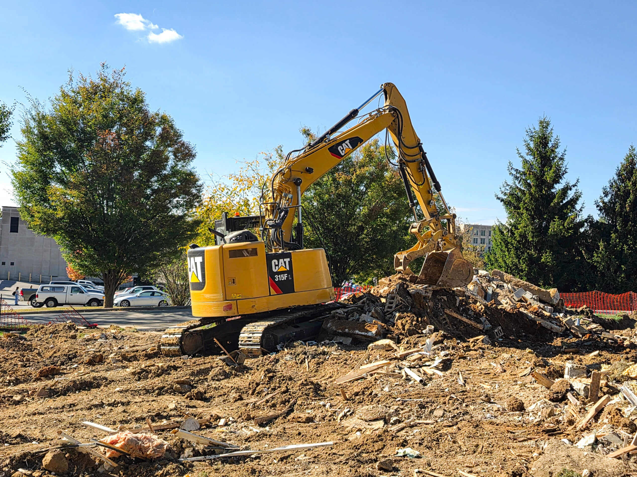 Elite Dumpster and Demolition Odon, IN Yellow CAT excavator clearing debris and rubble from a construction site, with trees and parked vehicles in the background under a clear sky.