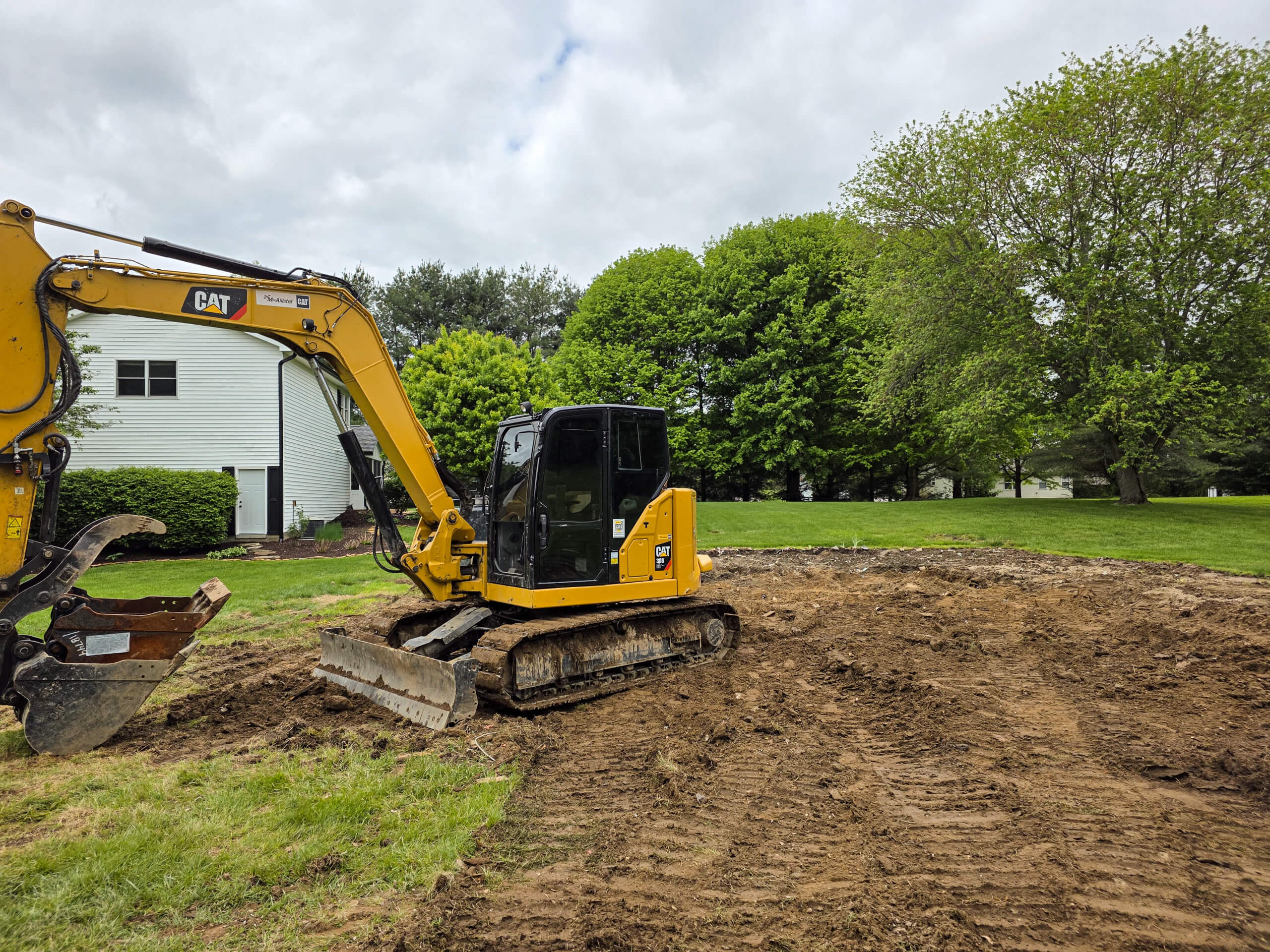 Elite Dumpster and Demolition Odon, IN A yellow excavator is parked on a patch of freshly dug earth in a grassy yard with trees and a white house in the background.
