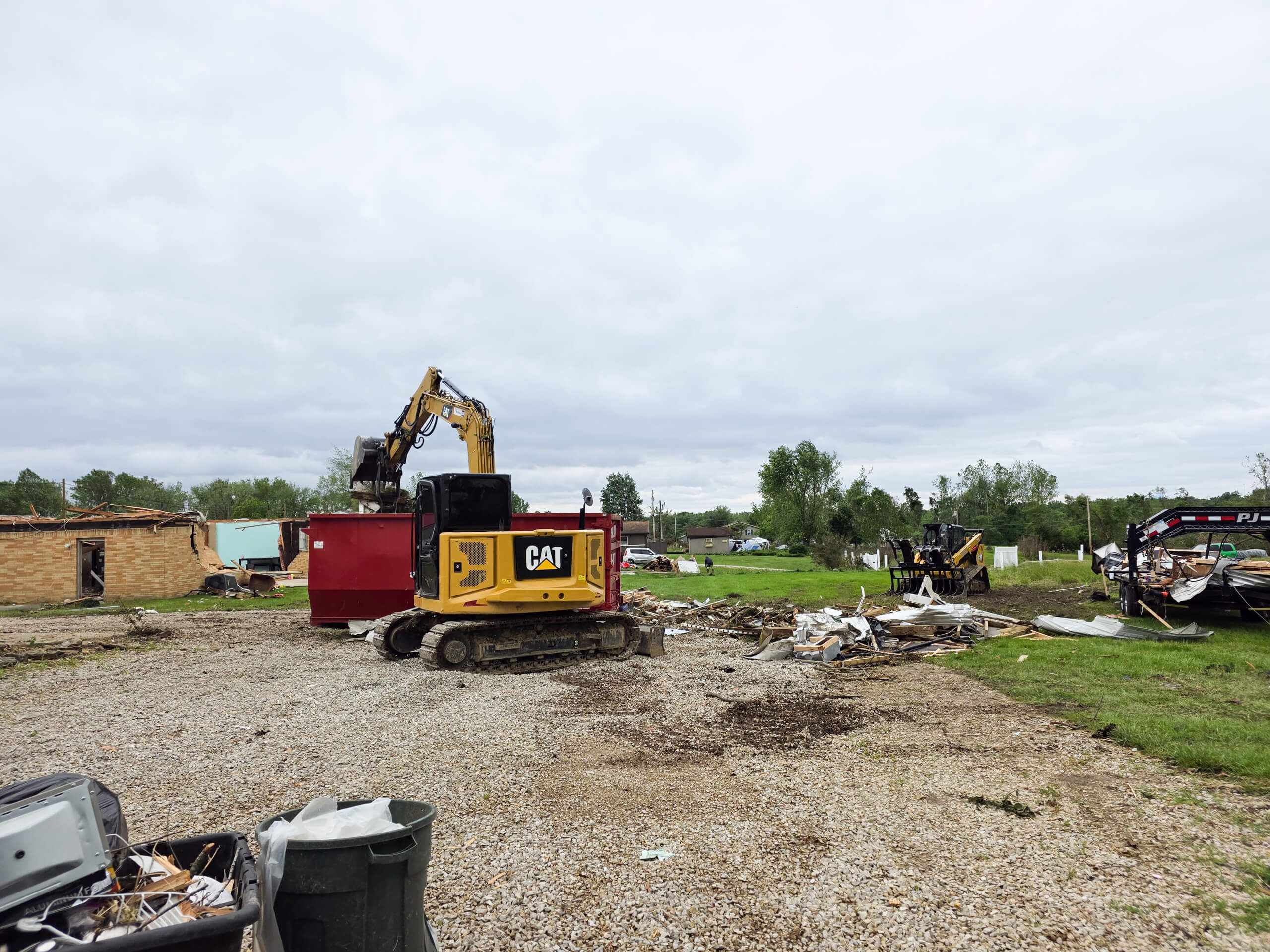 Elite Dumpster and Demolition Odon, IN A yellow excavator loads debris into a red dumpster at a demolition site, with scattered rubble and damaged structures visible in the background.