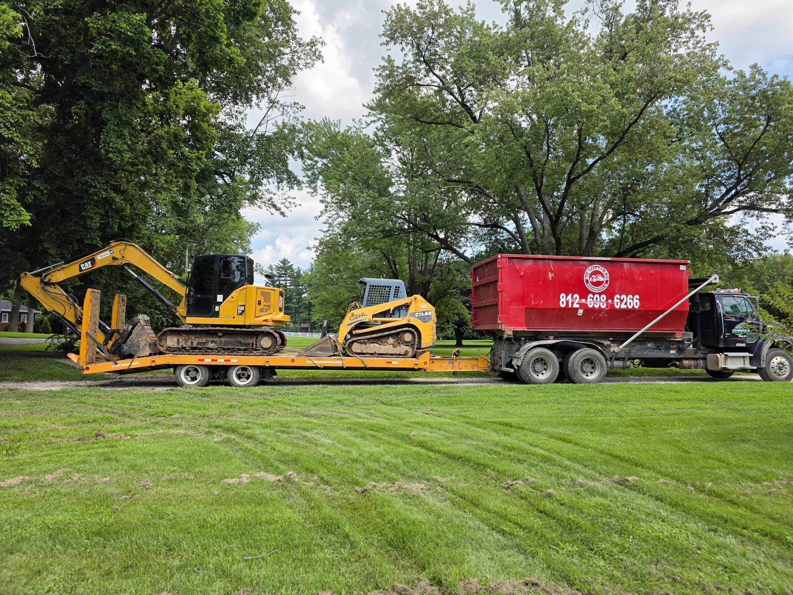 Elite Dumpster and Demolition Odon, IN A flatbed trailer carrying a yellow excavator and a compact track loader is parked next to a red dumpster bin truck on a grassy area with trees in the background.