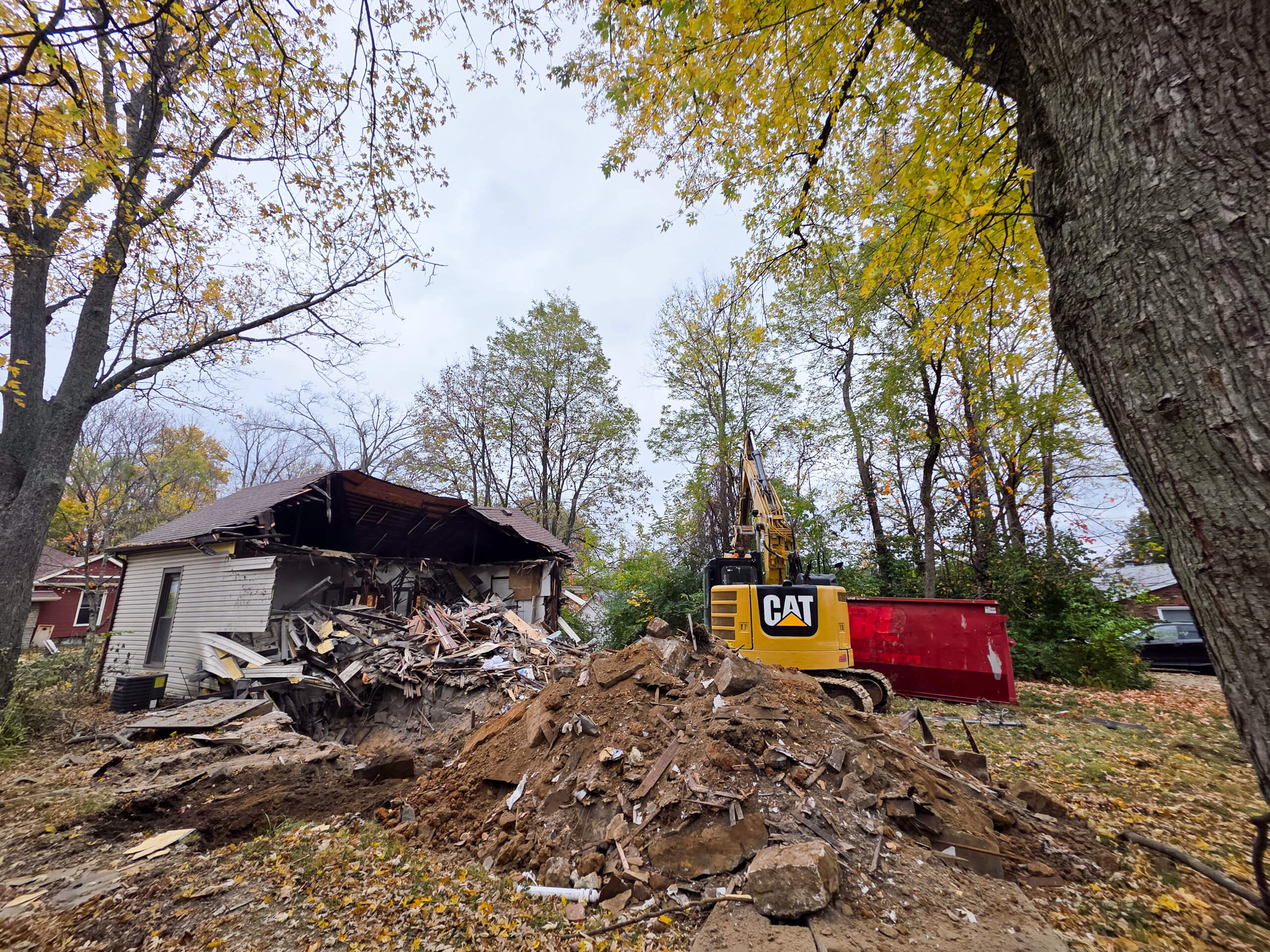Elite Dumpster and Demolition Odon, IN A partially demolished house with debris in front, a yellow excavator labeled "CAT," and trees with autumn leaves surrounding the scene.