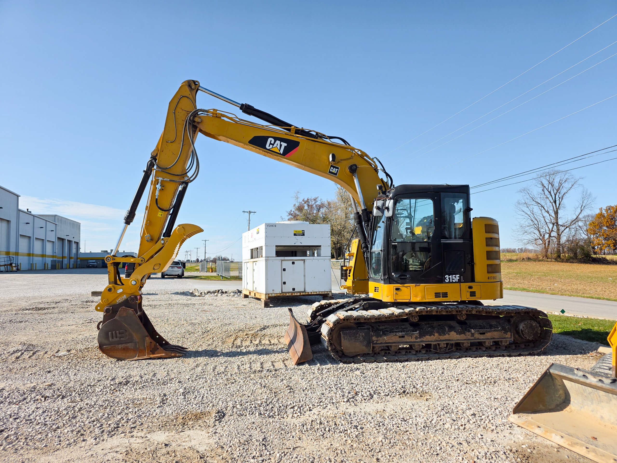 Elite Dumpster and Demolition Odon, IN A yellow CAT excavator is parked on a gravel lot near industrial buildings and a white generator unit under a clear blue sky.