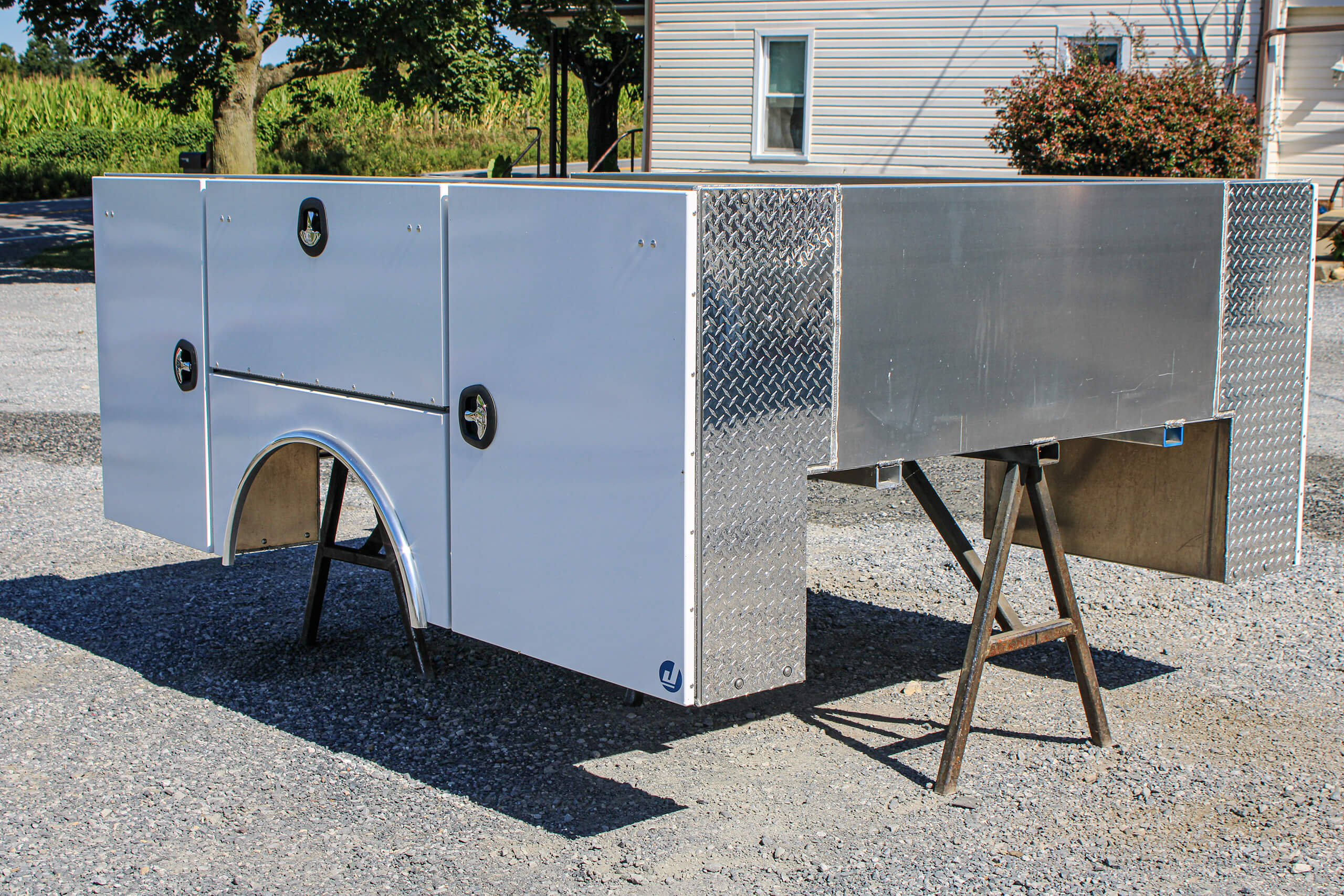 Metal utility truck bed with side storage compartments is positioned on two sawhorses outdoors near a building and a tree.