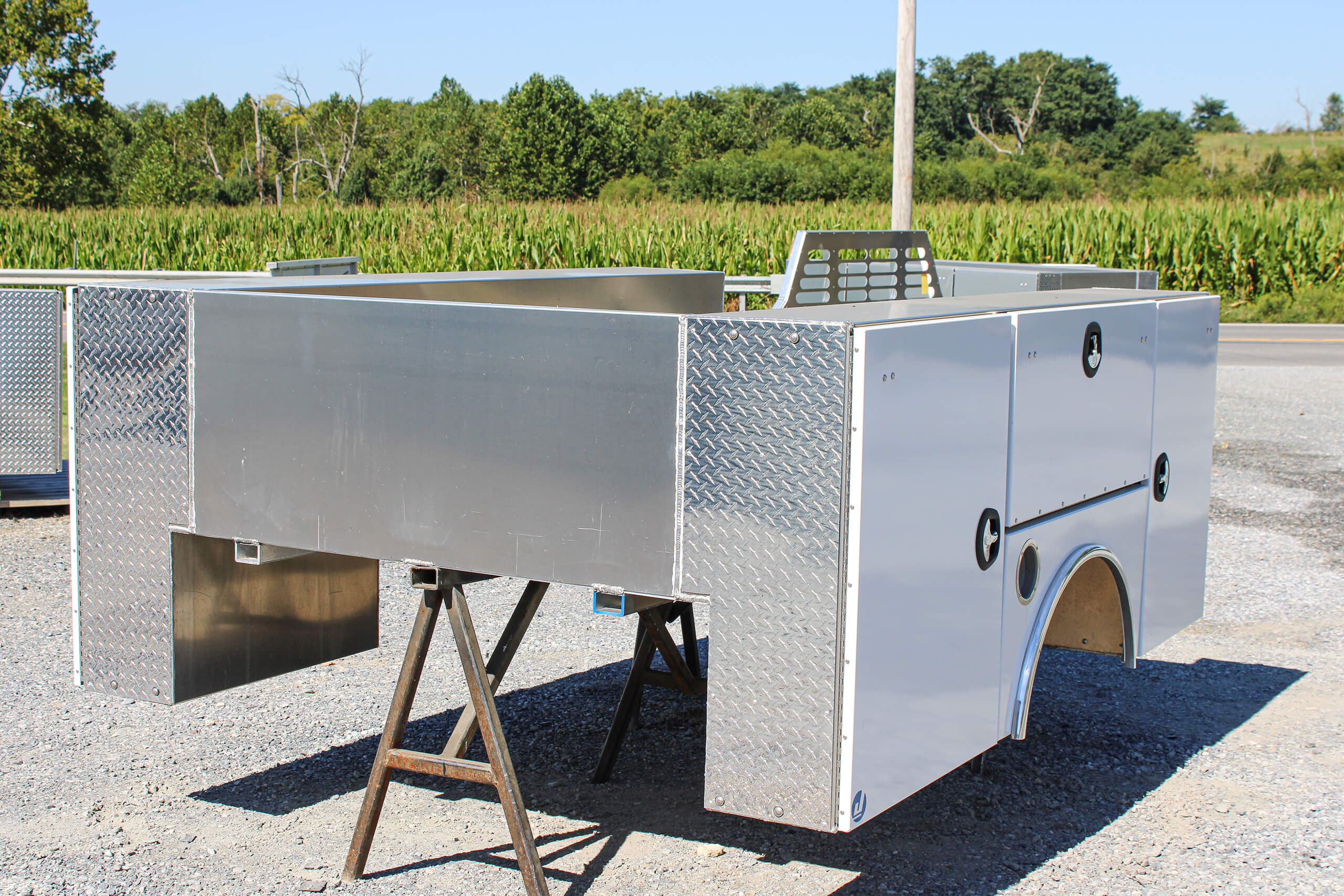 A metallic utility truck bed with storage compartments is propped up on two wooden sawhorses outdoors near a road and greenery.