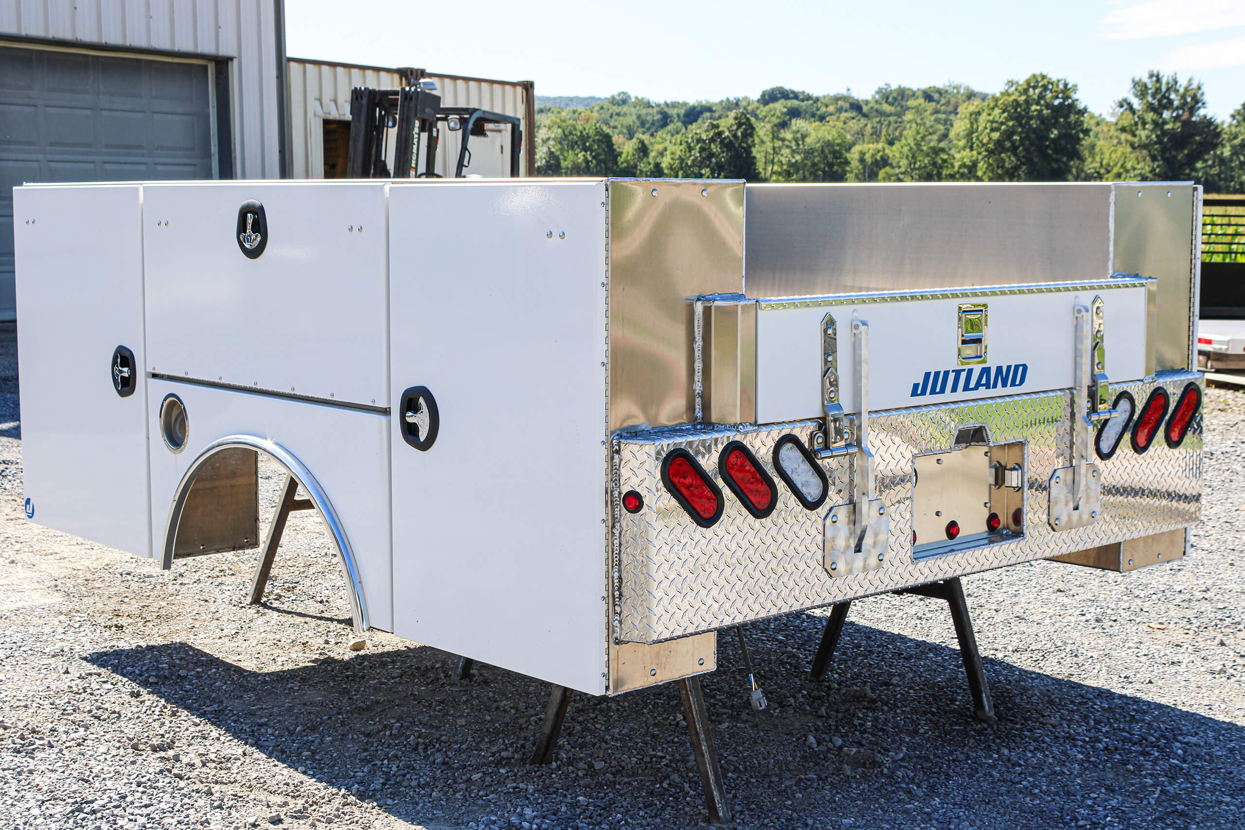 A white utility truck bed with storage compartments and rear light clusters is elevated on stands in an outdoor lot near a building.