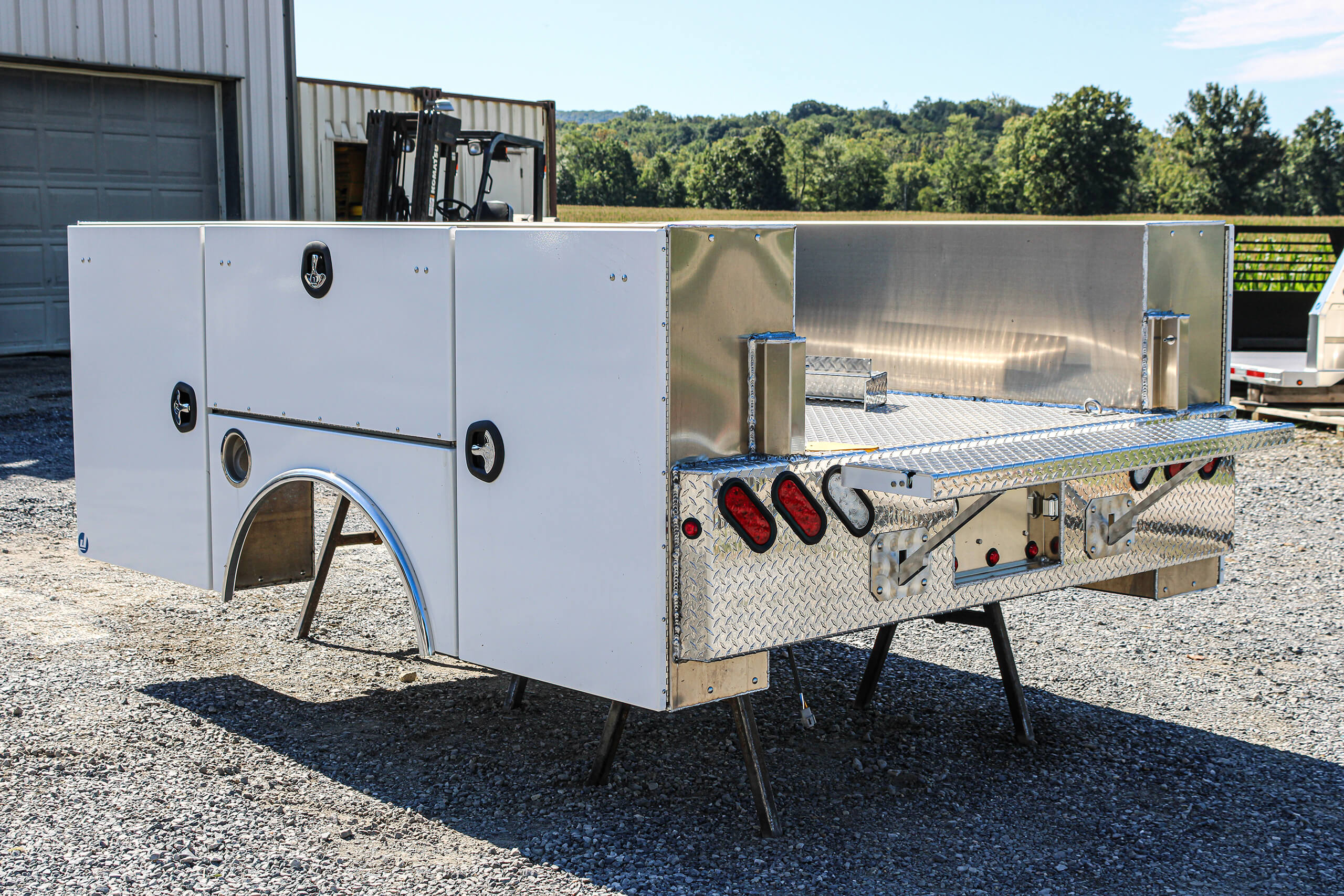 White utility truck bed with metal storage compartments and rear taillights, displayed outdoors on stands near a building and trees.