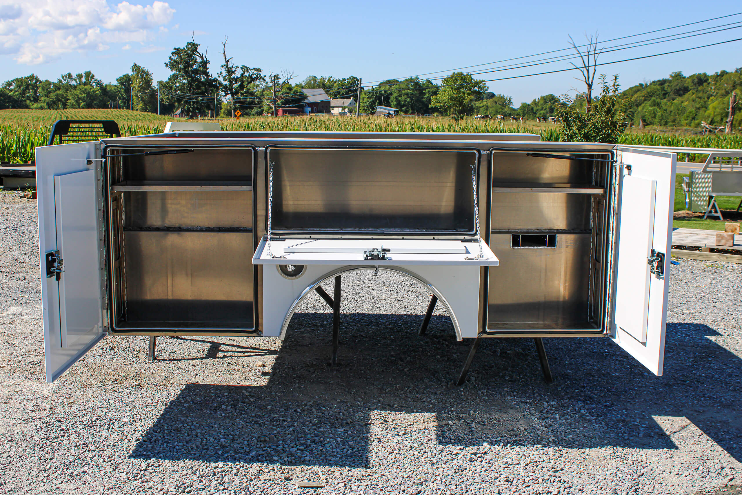A white metal utility truck bed with both side compartments and rear doors open, revealing empty storage spaces, is parked on gravel with a field and trees in the background.