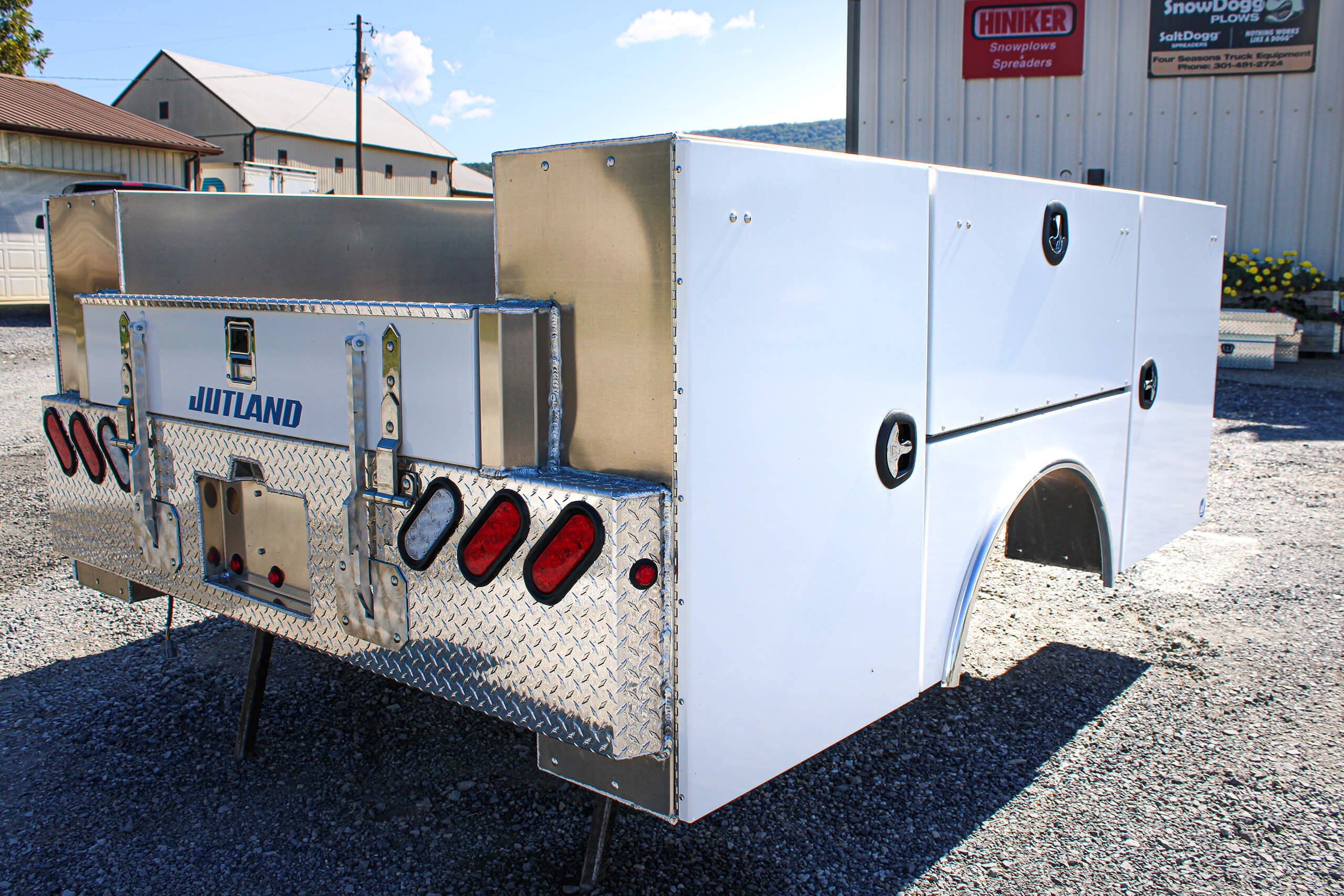 A white utility truck bed with diamond plate accents and multiple storage compartments is displayed outdoors on gravel, near a building.