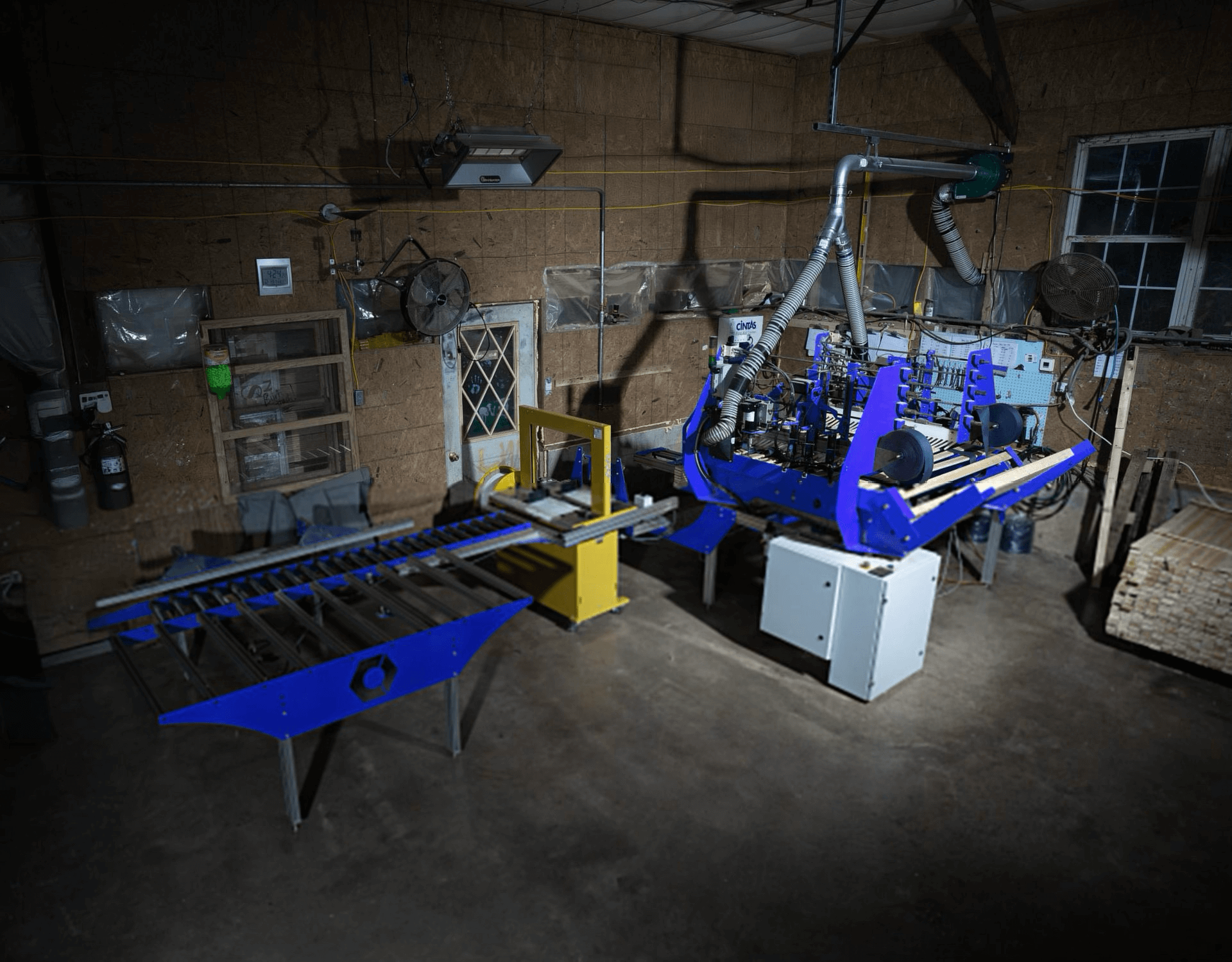 A blue and silver industrial machine with rollers is set up in a workshop with concrete floors, wooden walls, and scattered equipment.