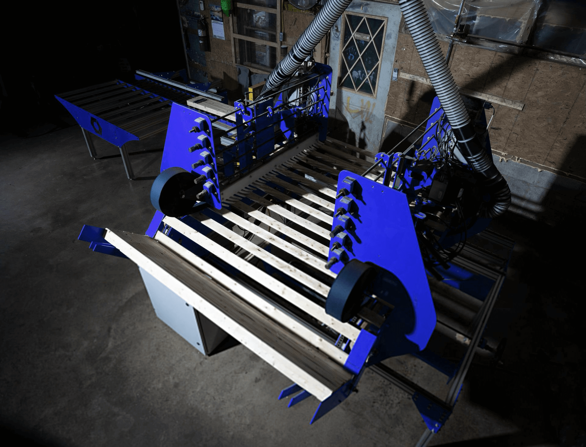 A large industrial machine with blue components processes wooden planks inside a workshop with unfinished walls.