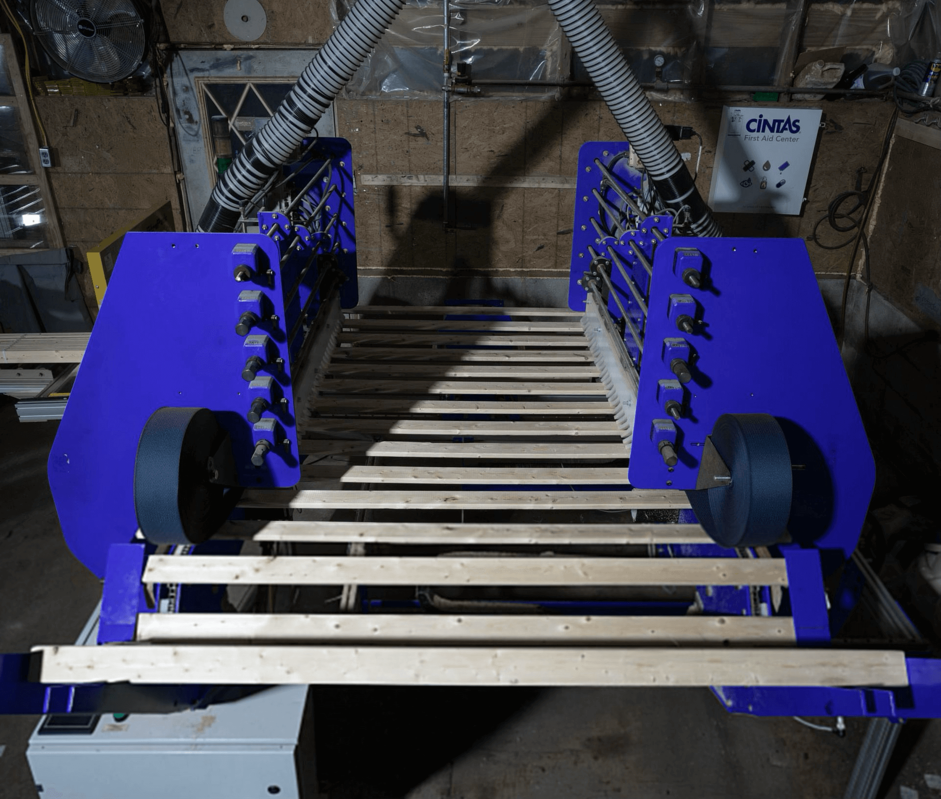 A large blue industrial machine with rollers and ventilation hoses holds several wooden planks inside a workshop.