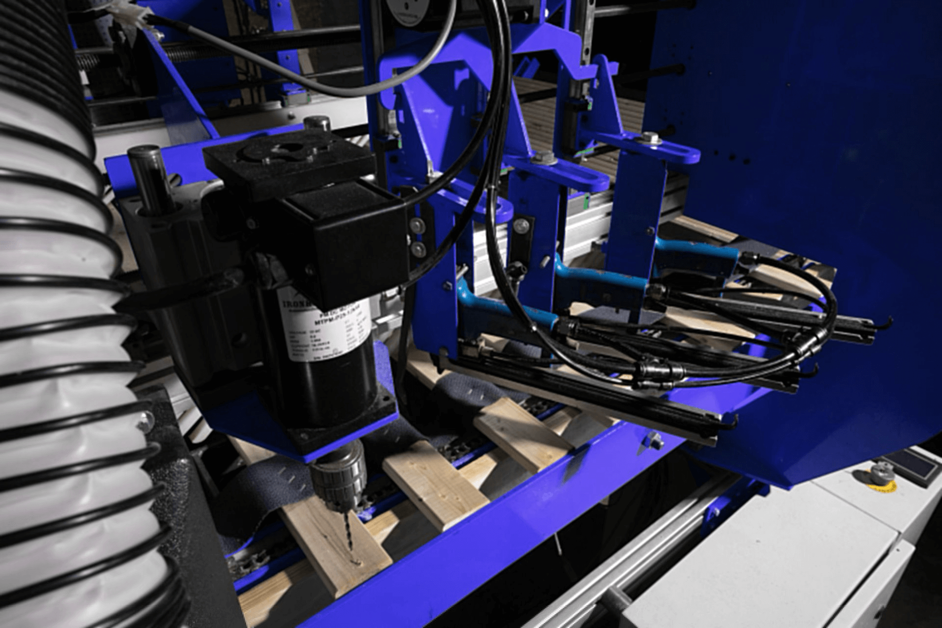 Automated industrial machine with a drill head working on wooden planks, surrounded by blue structural components and cables.