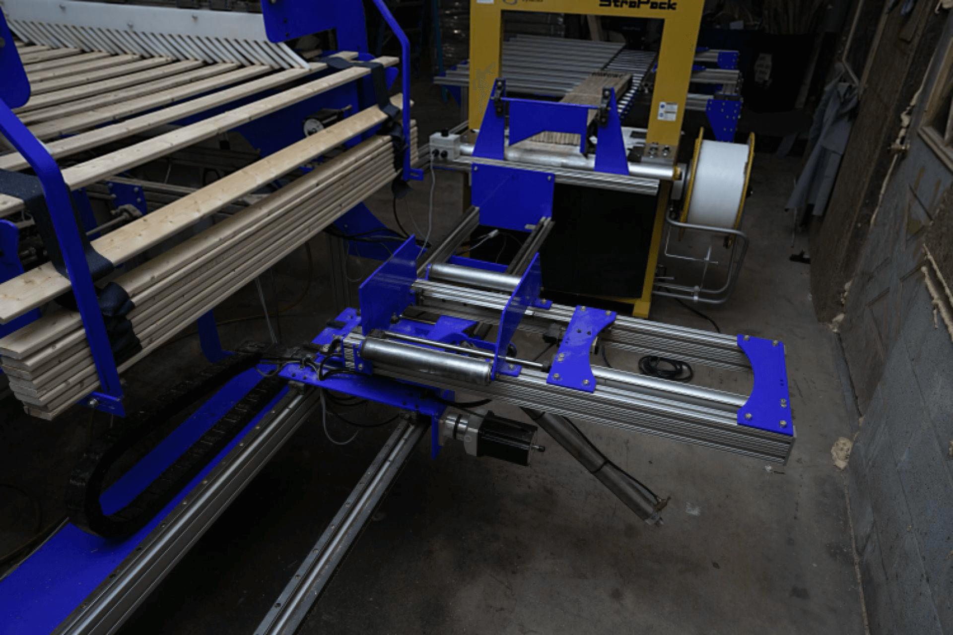 A blue and silver automated industrial machine with metal rollers and wood planks, located in a warehouse setting.