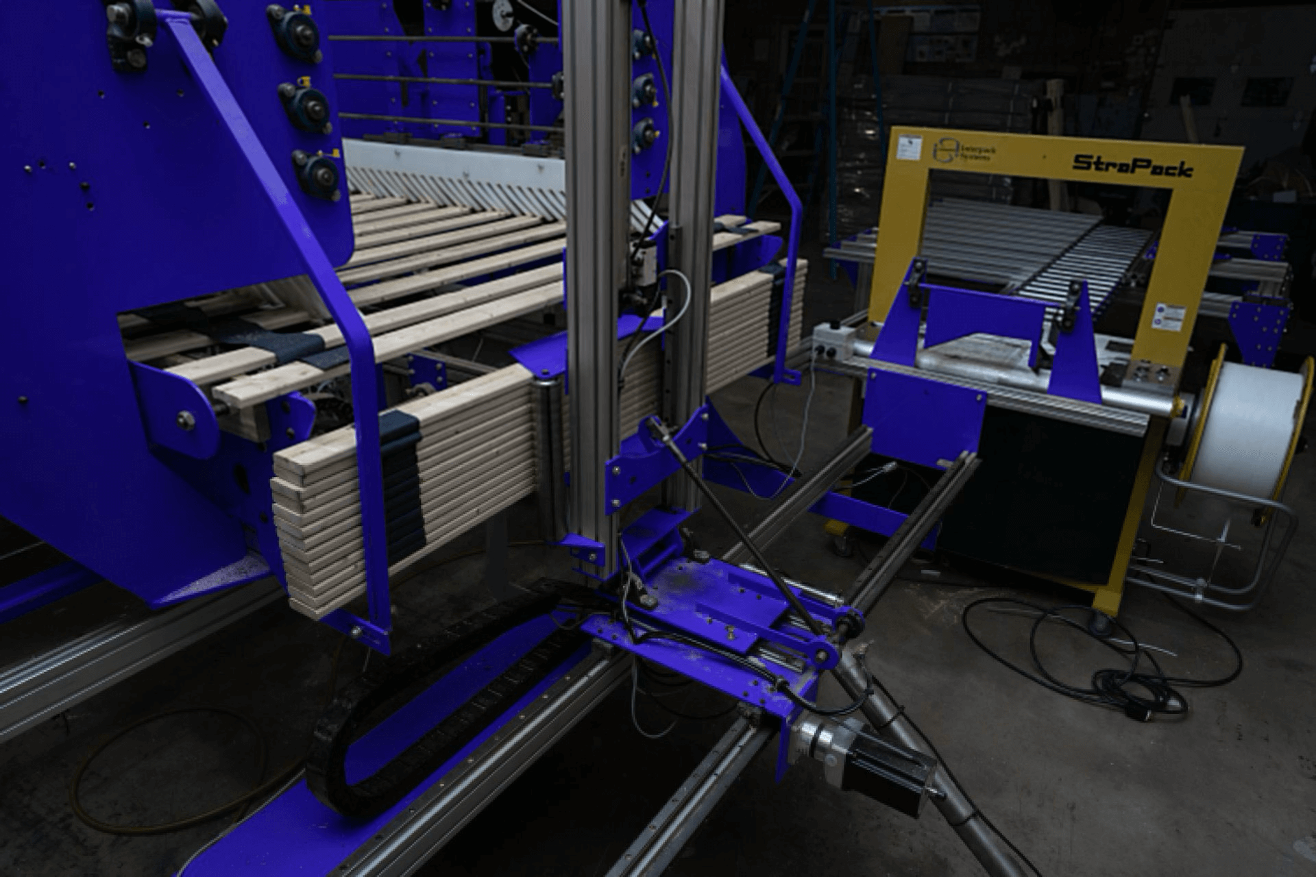 A packaging machine with stacked wooden planks is positioned next to a yellow StraPack strapping machine in an industrial setting.