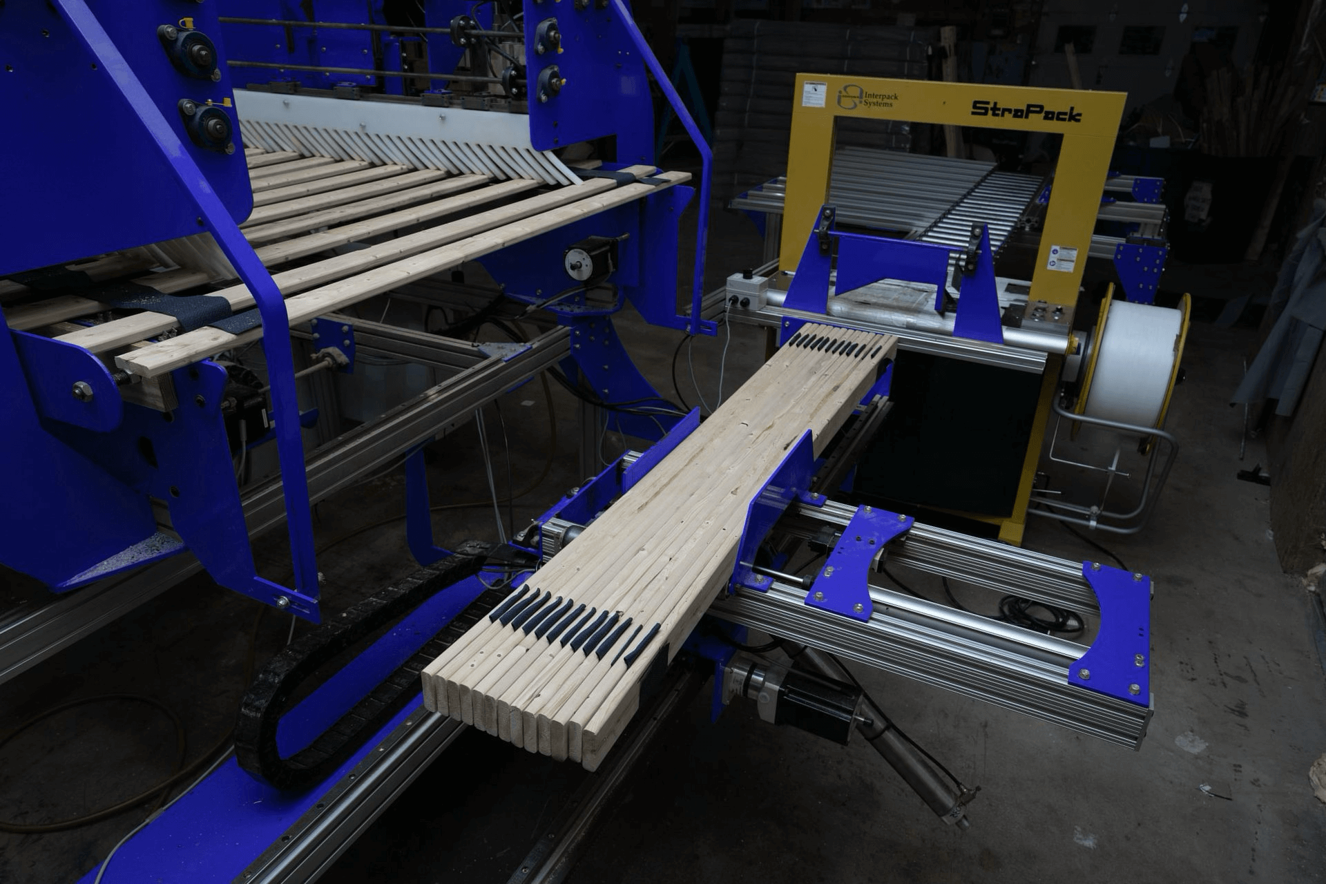 Wooden slats are being processed on an automated assembly line with blue and yellow industrial machinery in a factory setting.