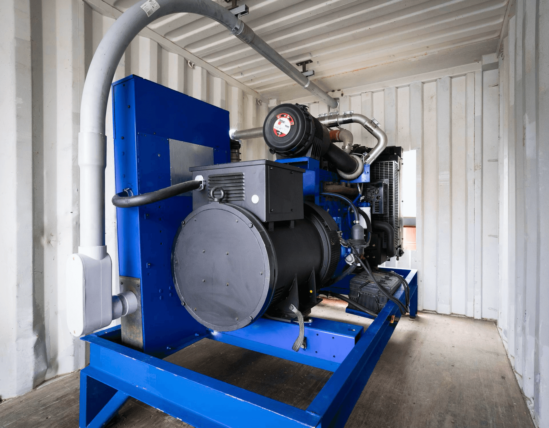A large blue industrial generator is installed inside a white metal shipping container with visible wiring and ventilation pipes.