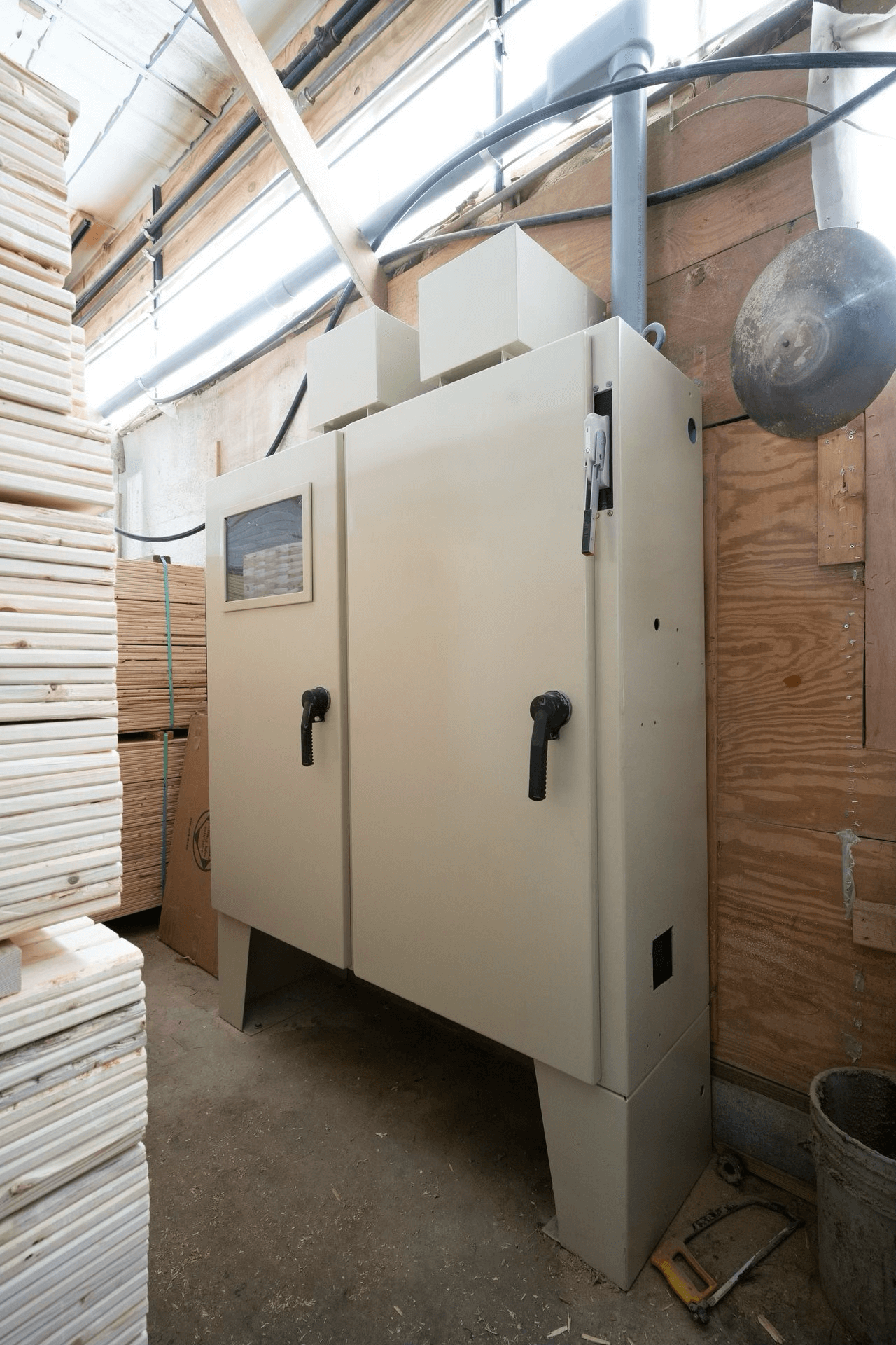 Large industrial electrical control panel with two doors and handles, installed indoors, surrounded by stacked wood panels and exposed wiring.