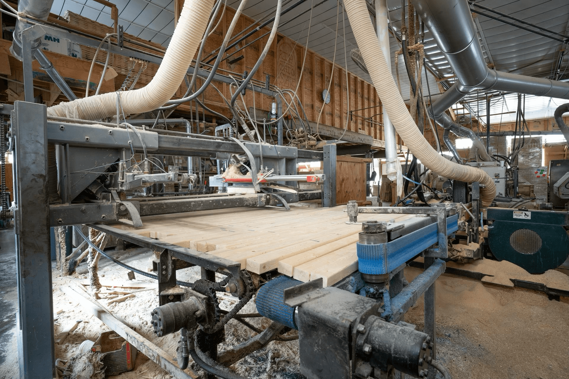 Large industrial woodworking machine in a factory processes wooden boards, surrounded by sawdust, pipes, and various mechanical components.