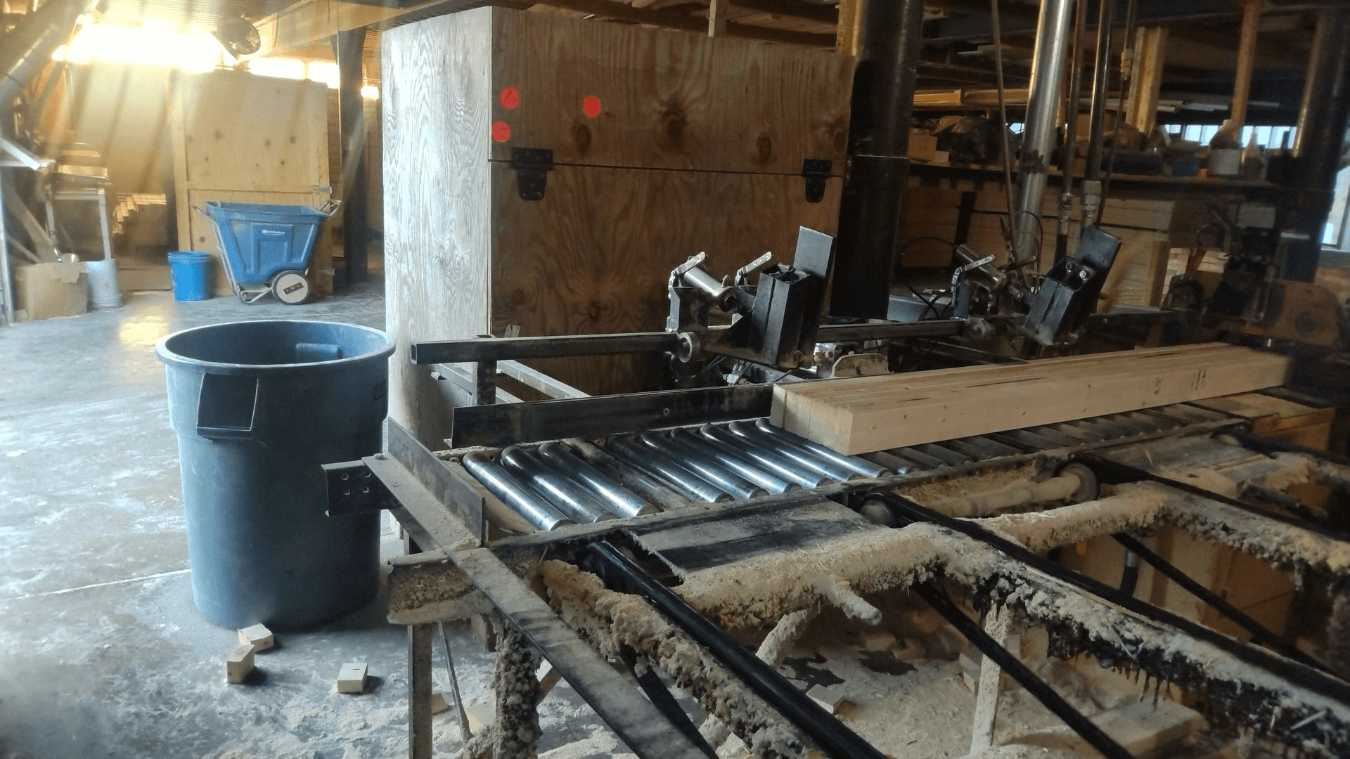 A piece of lumber on a roller conveyor in a woodworking facility, surrounded by sawdust, industrial machinery, and storage containers.