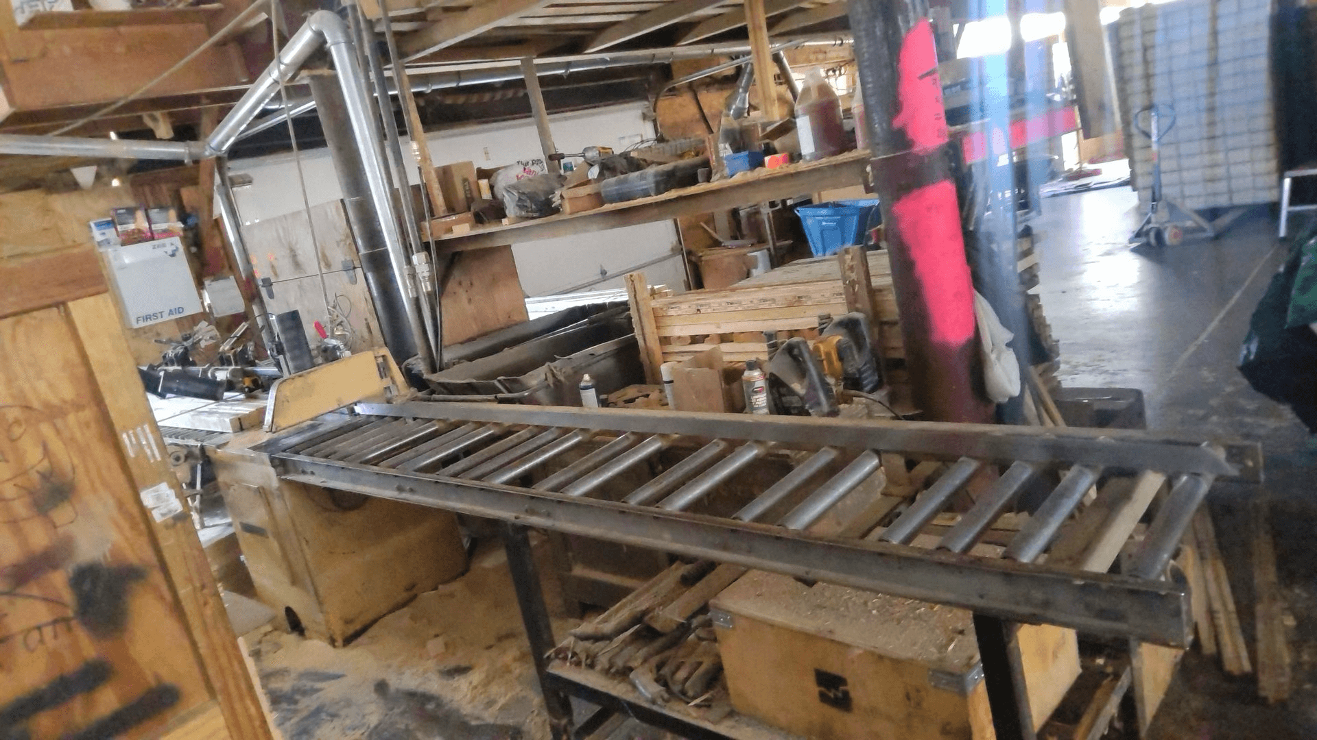 A metal roller conveyor in a cluttered workshop with wooden shelves, assorted tools, and equipment in the background.
