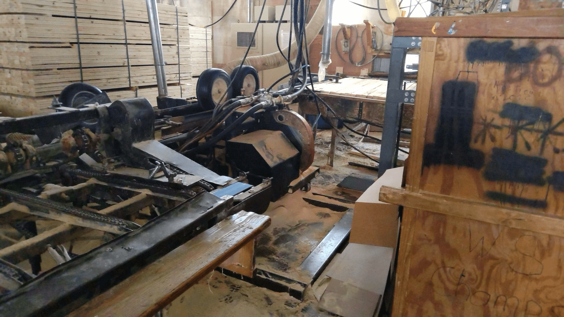 A cluttered workshop with industrial machinery, wooden crates, stacked boards, cables, and sawdust covering the floor.