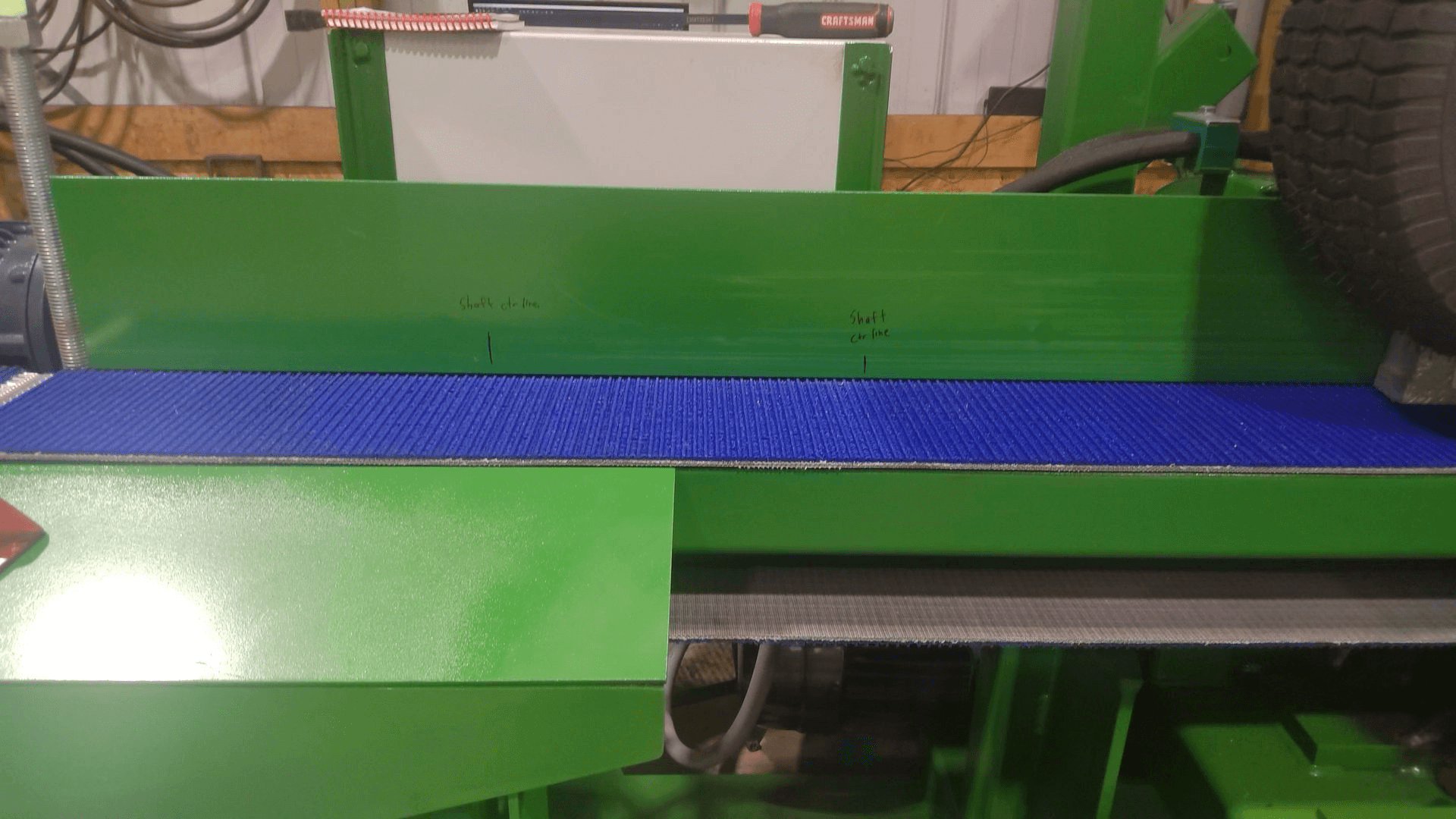 A green industrial machine with a blue textured conveyor belt, featuring handwritten measurements on the green surface above the belt.