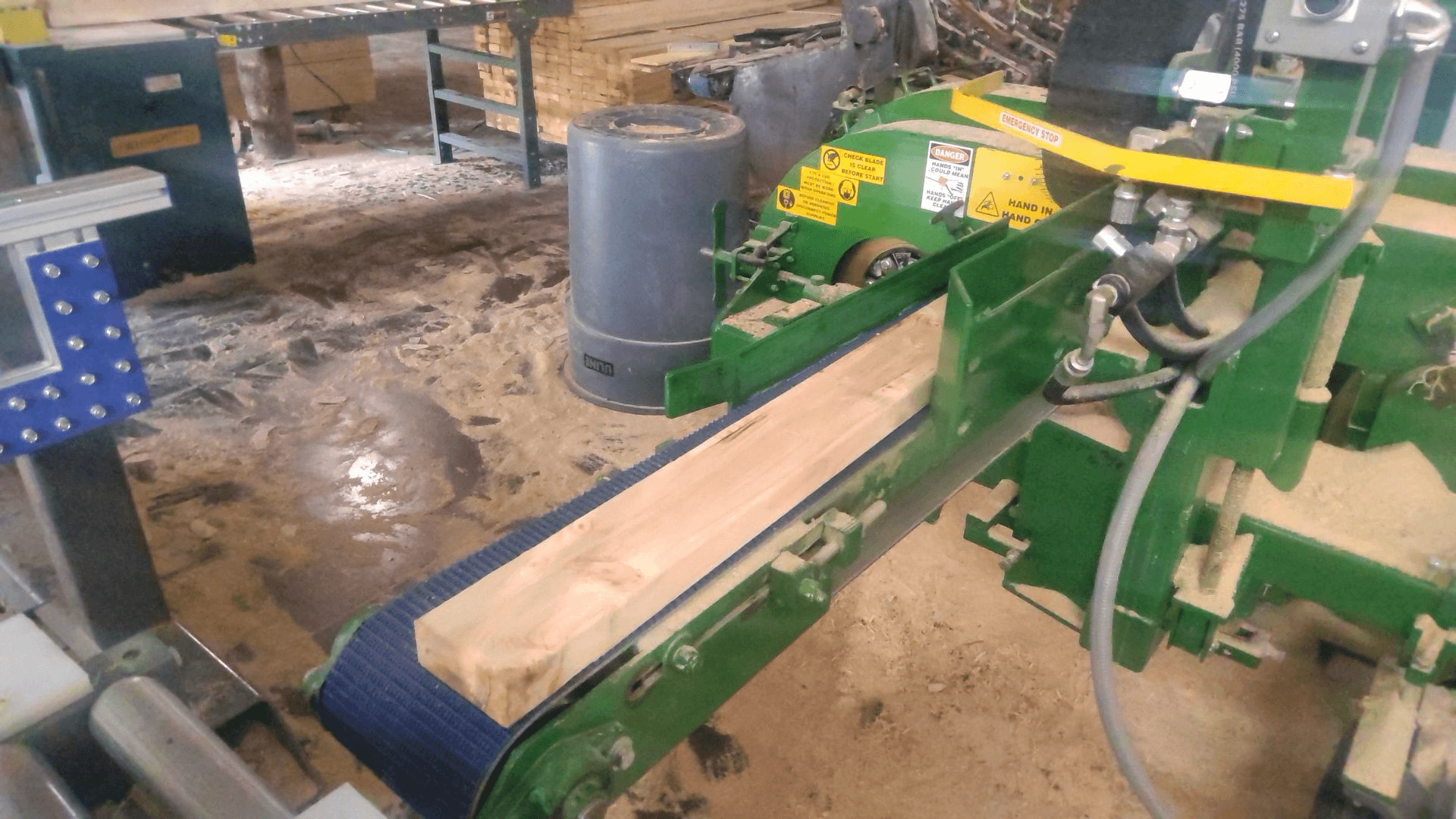 A piece of wood moves along a green conveyor belt in a sawmill with machinery, sawdust, and equipment visible in the background.