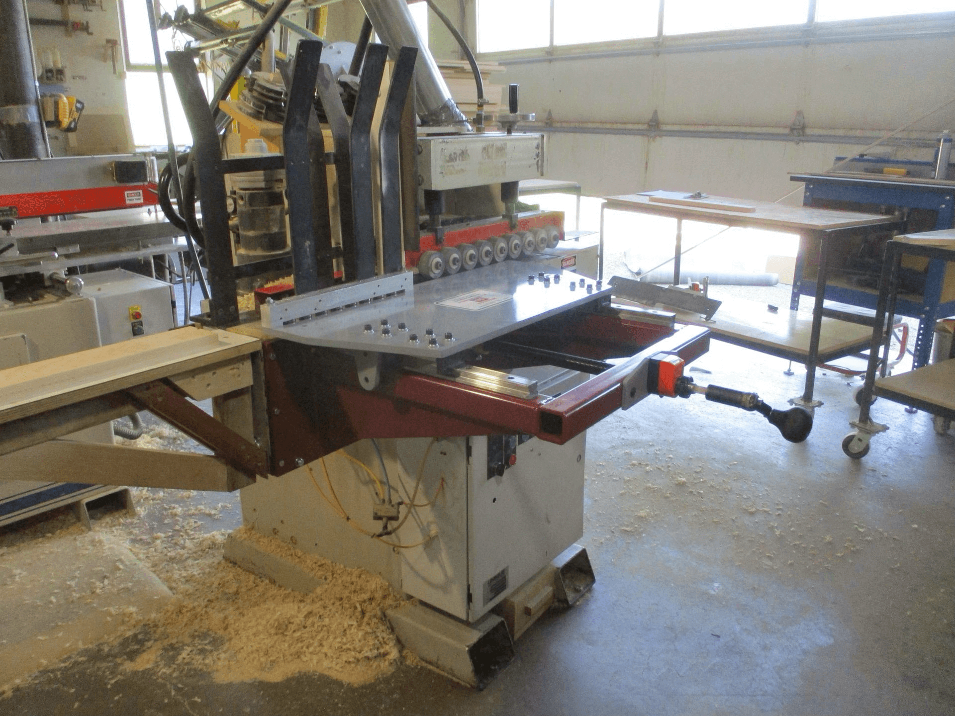 Industrial woodworking machine with metal clamps and cutting blades, surrounded by wood shavings in a workshop setting.