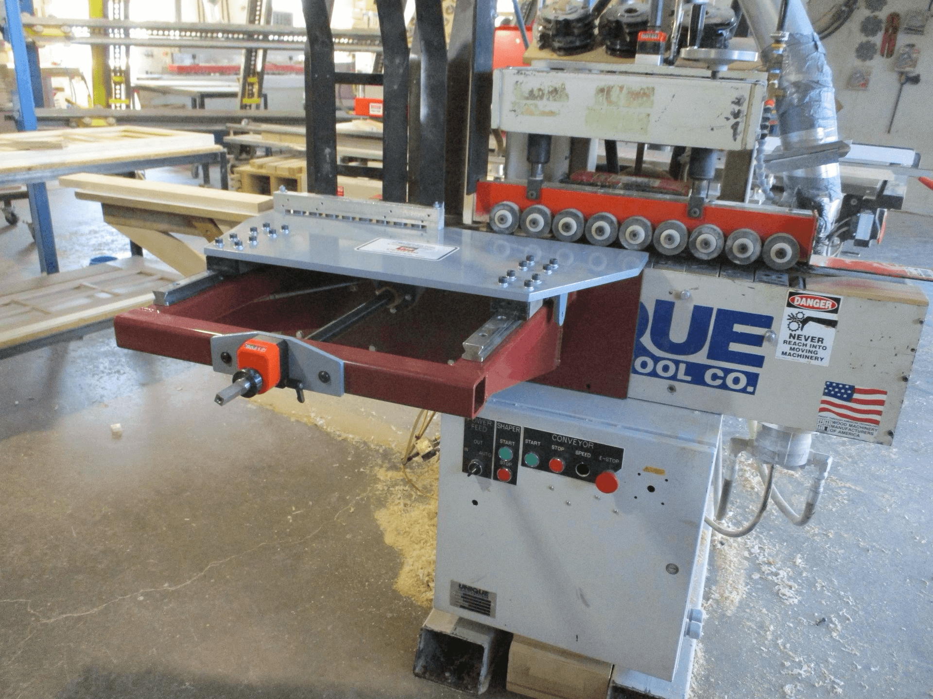 Industrial woodworking machine with control panel and warning labels in a workshop, dust and wood shavings on floor, shelving and equipment in the background.