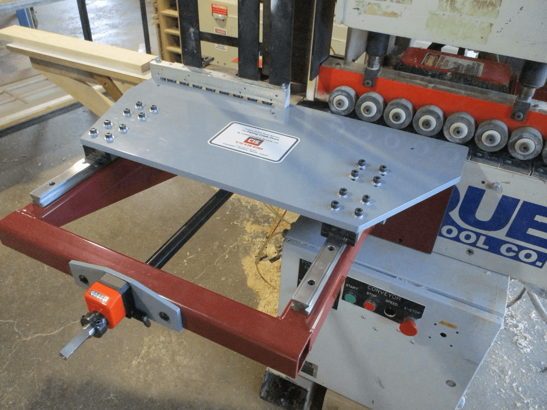 A metal jig fixture is attached to a woodworking machine in a workshop, with sawdust scattered on the floor.
