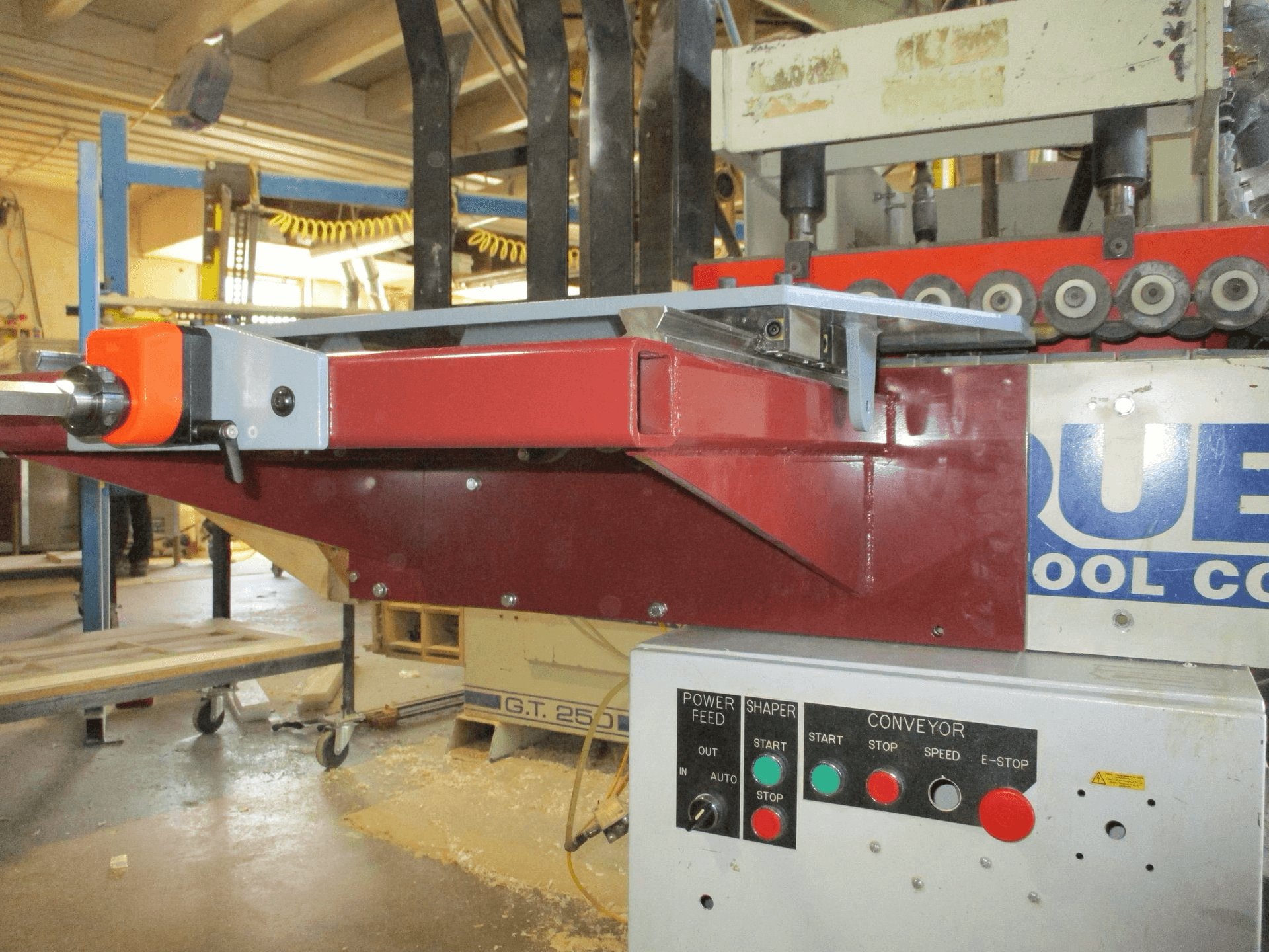 A close-up view of industrial woodworking machinery with visible control panel, buttons, and conveyor system in a workshop setting.