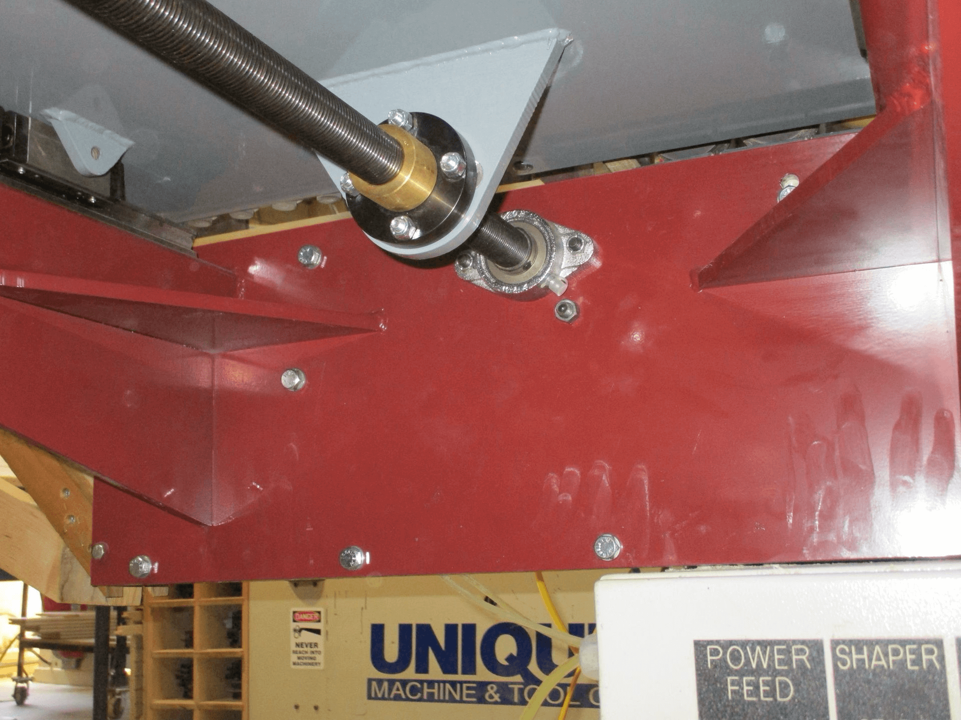 Close-up view of a machine component with a metal shaft, bearings, and red metal frame. The background shows part of another machine labeled 