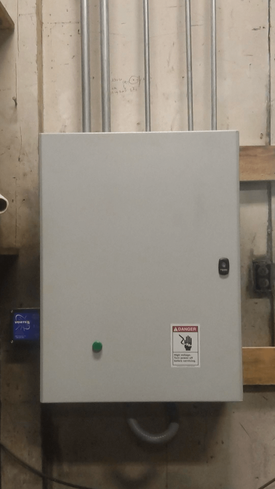 A gray electrical control panel with visible conduit pipes above, a green button, and a danger warning label is mounted on a wall in an industrial setting.