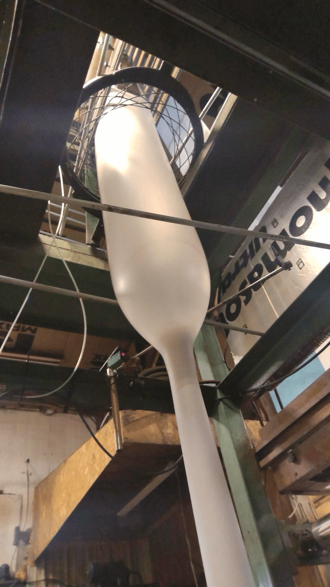 A large tube of white plastic is being extruded vertically upwards in an industrial setting with metal platforms and machinery.