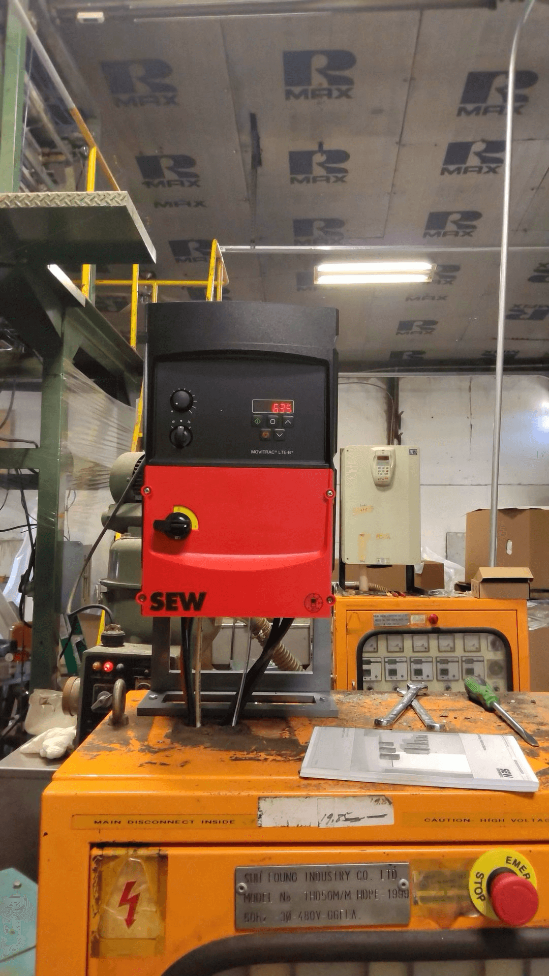 An SEW-branded industrial control unit sits atop an orange machine in a factory setting, with a digital display showing “6.25.” Boxes and electrical panels are visible in the background.