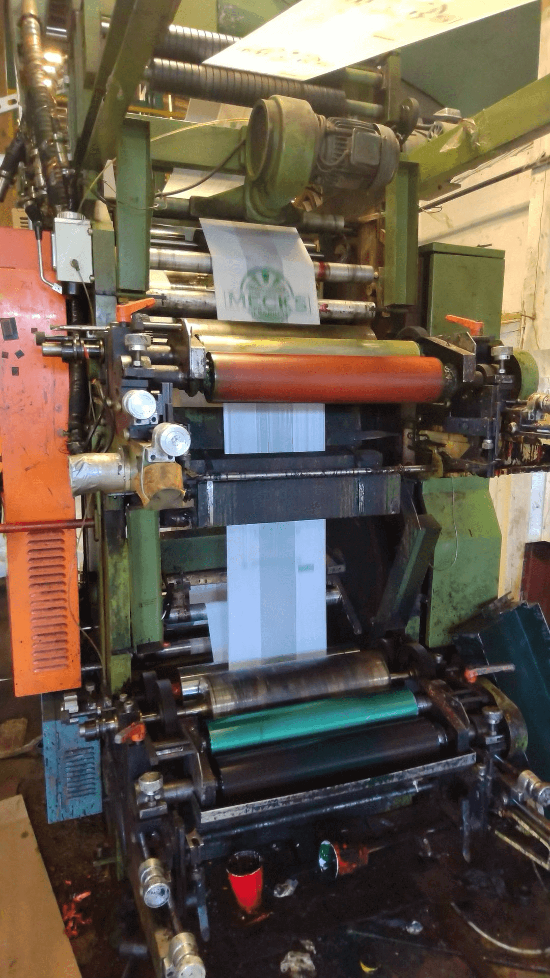A large, industrial printing press in operation, producing printed sheets with green ink on a white material. Various rollers, gears, and mechanical parts are visible.