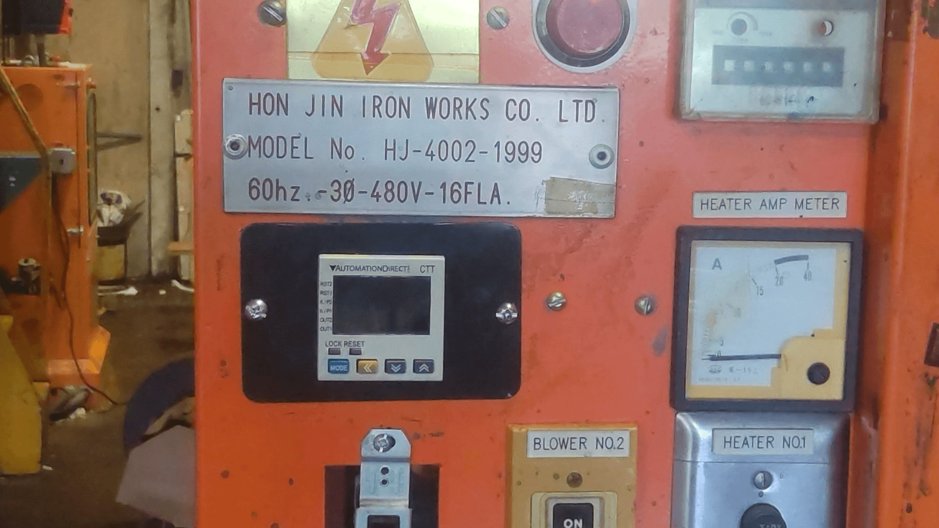 Close-up of an orange industrial machine panel showing meters, switches, and a label from Hon Jin Iron Works Co. Ltd. with model and electrical details.
