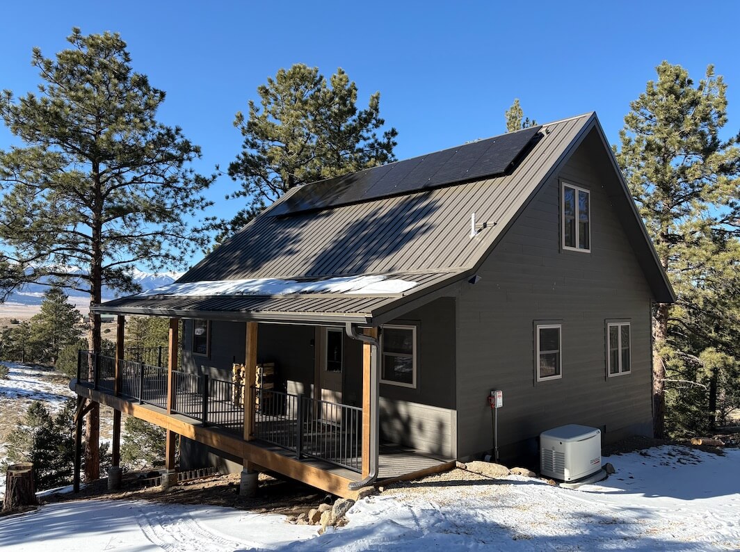 A gray cabin with solar panels on the roof is surrounded by pine trees and patches of snow, with a large porch and a generator visible near the building.