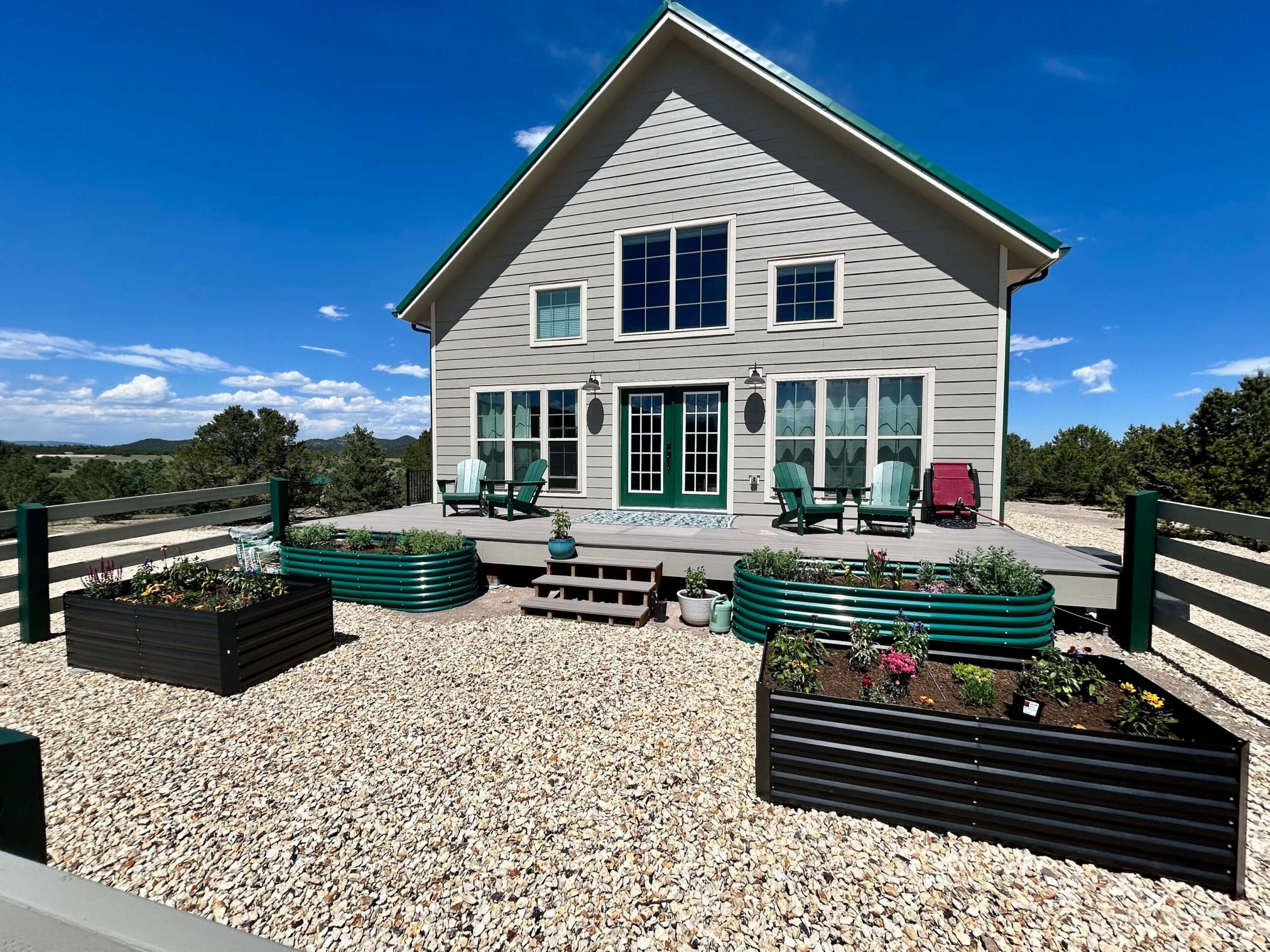 A modern house with large windows, a deck with outdoor seating, and raised metal garden beds on a gravel-covered yard under a clear blue sky.