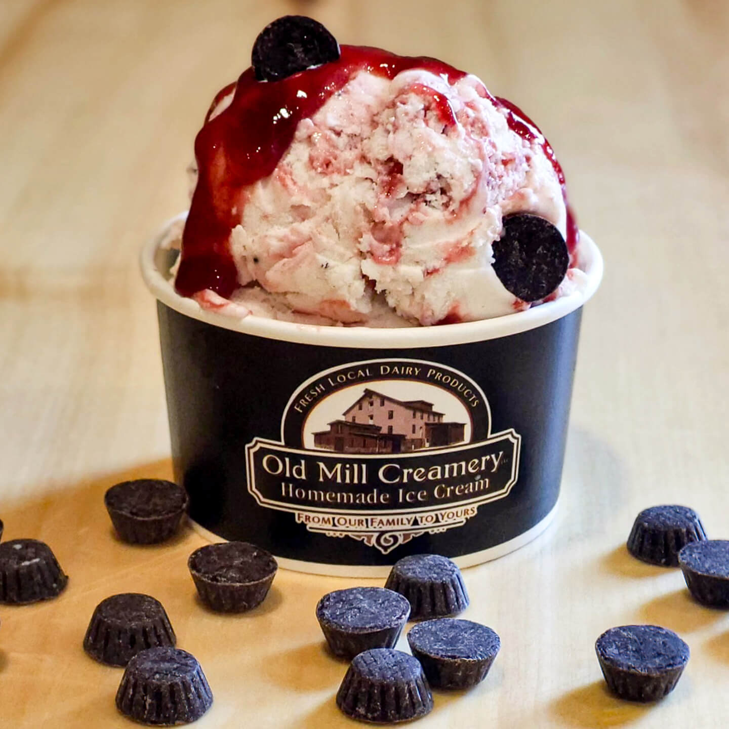 A cup of Old Mill Creamery ice cream topped with berry sauce and chocolate pieces, with more chocolate pieces scattered on a light wooden surface.