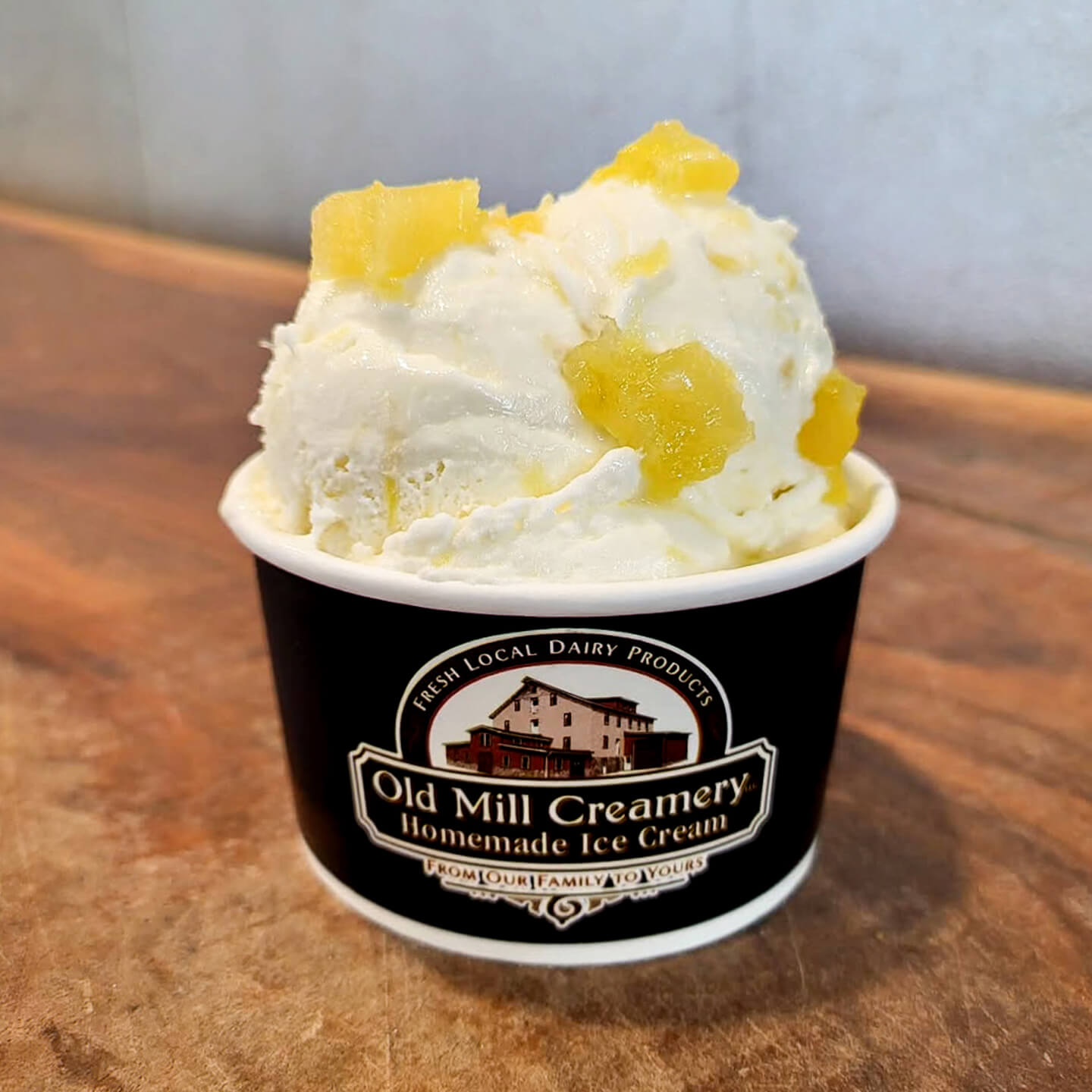 A cup of vanilla ice cream with pineapple chunks in a black Old Mill Creamery branded container on a wooden surface.