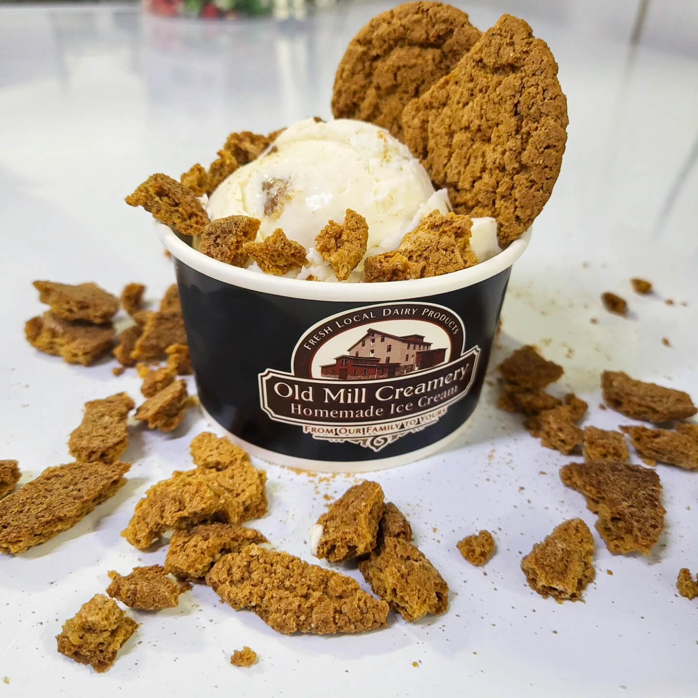 A cup of vanilla ice cream topped with two large cookies, surrounded by broken cookie pieces on a white surface. The cup is labeled “Old Mill Creamery Homemade Ice Cream.”.