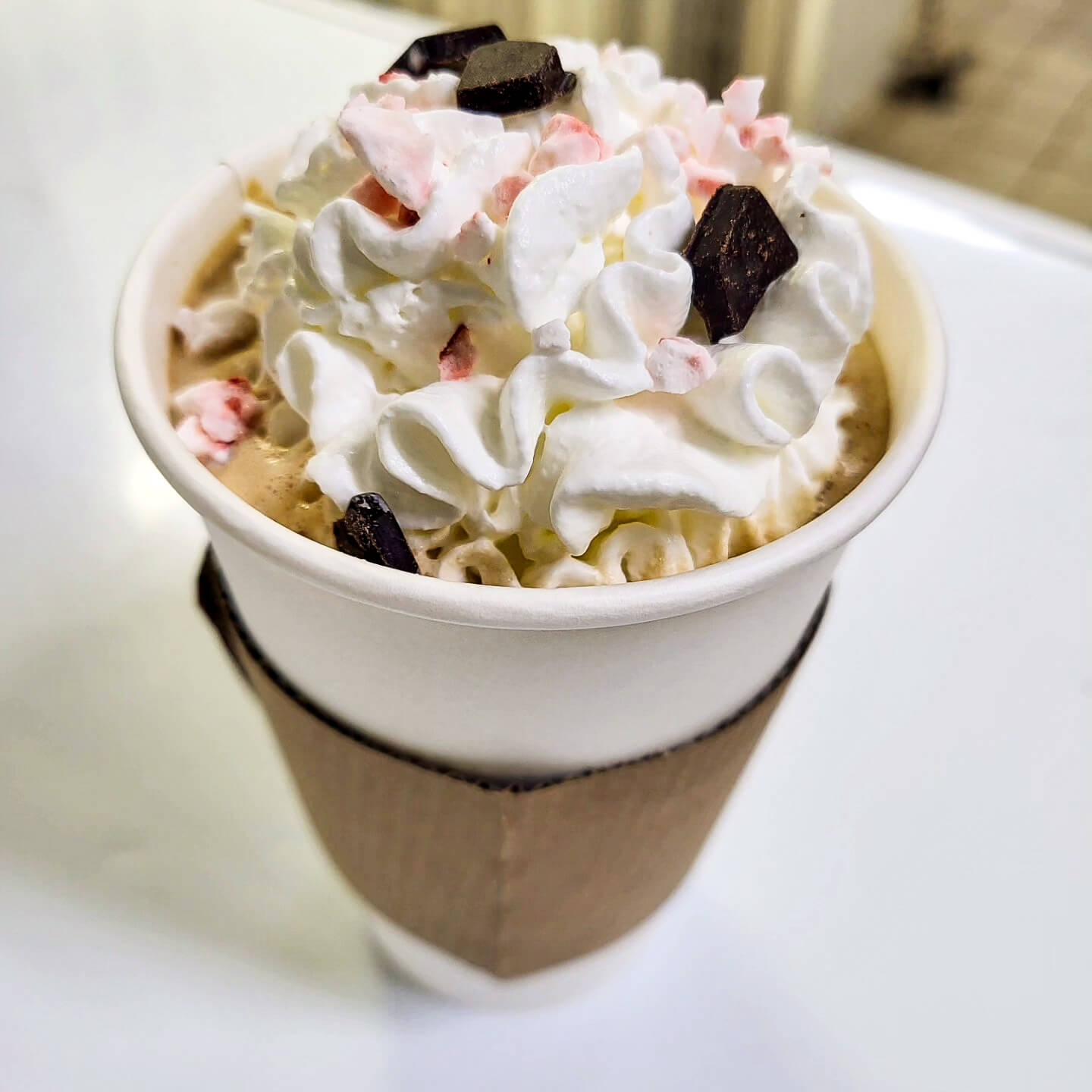 A hot beverage in a white paper cup topped with whipped cream, chocolate chunks, and pink peppermint pieces, with a cardboard sleeve around the cup.