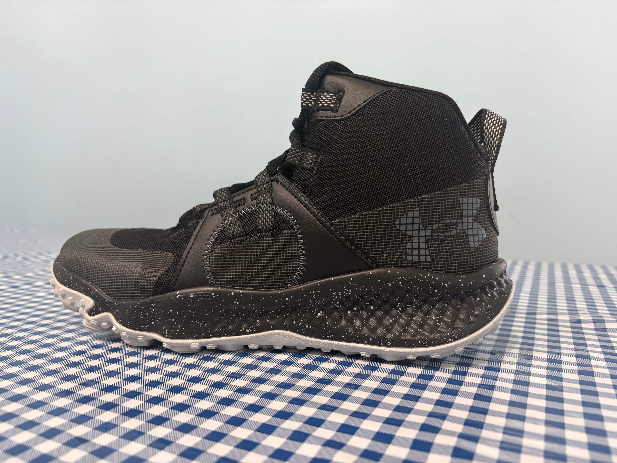 3.14.26 Miller's Country Store Under Armor Shoe Ad A black Under Armour high-top sneaker with a textured sole sits on a blue and white checkered surface.