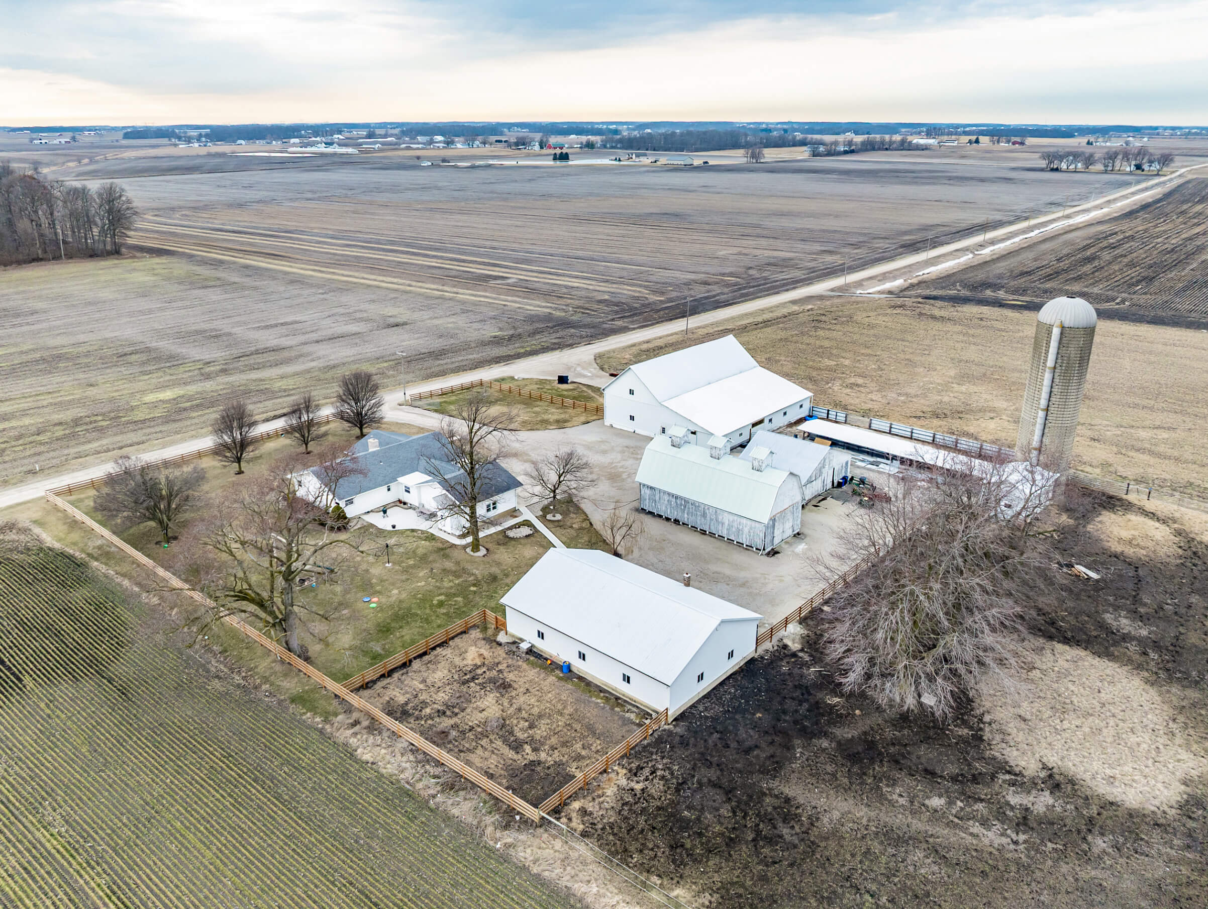 Cardinal Commercial Roofing Decatur IN Aerial view of a farm with several white buildings, a silo, fenced yard, and surrounding fields in a rural landscape.
