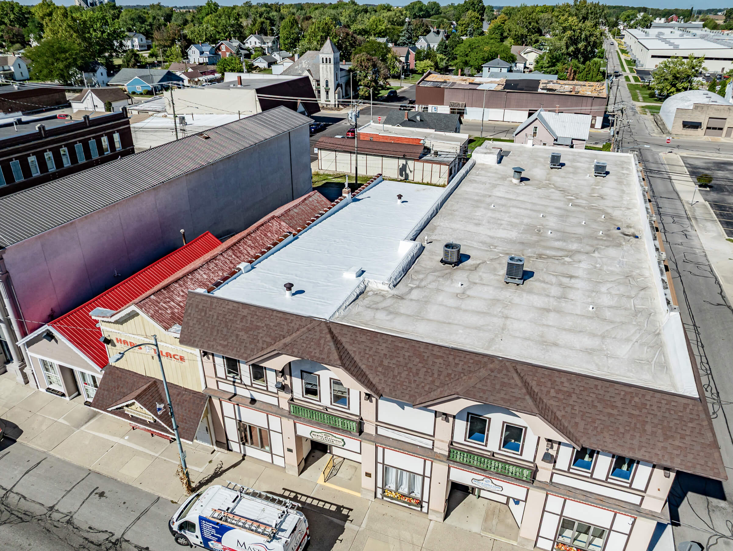 Cardinal Commercial Roofing Decatur IN Aerial view of a commercial building with a flat white roof and storefronts, next to a red-roofed structure, along a city street with a parked van and nearby residential area.