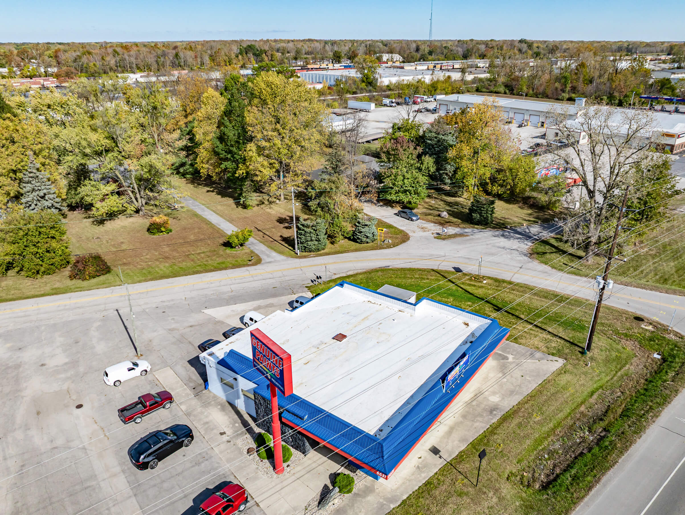 Cardinal Commercial Roofing Decatur IN Aerial view of a freestanding white-roofed commercial building with a red and blue exterior, parking lot, and cars, surrounded by trees and nearby roads in a suburban area.