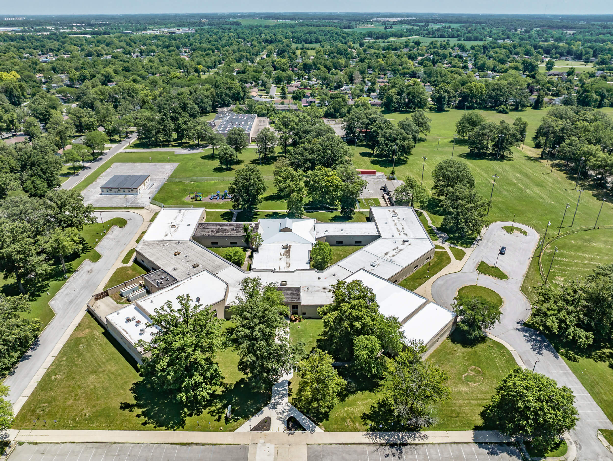 Cardinal Commercial Roofing Decatur IN Aerial view of a large, single-story school building with a white roof, surrounded by trees, parking lots, and open green fields in a suburban area.
