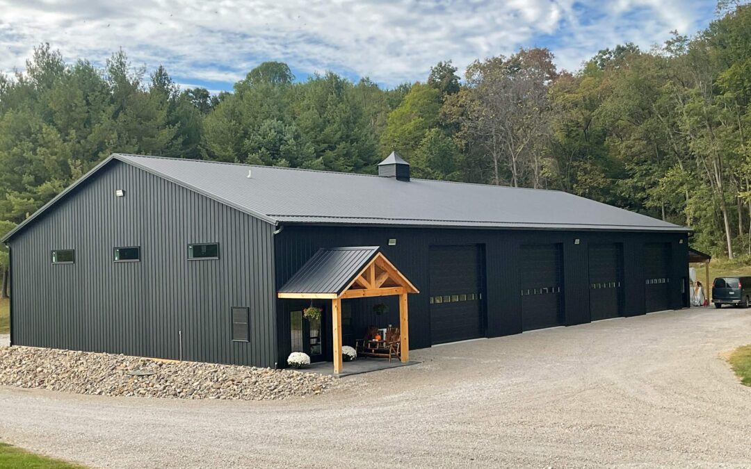 Amish Ridge Metals Mt Perry OH Large black metal building with four garage doors, a small wooden entrance, and a gravel driveway surrounded by trees and greenery.