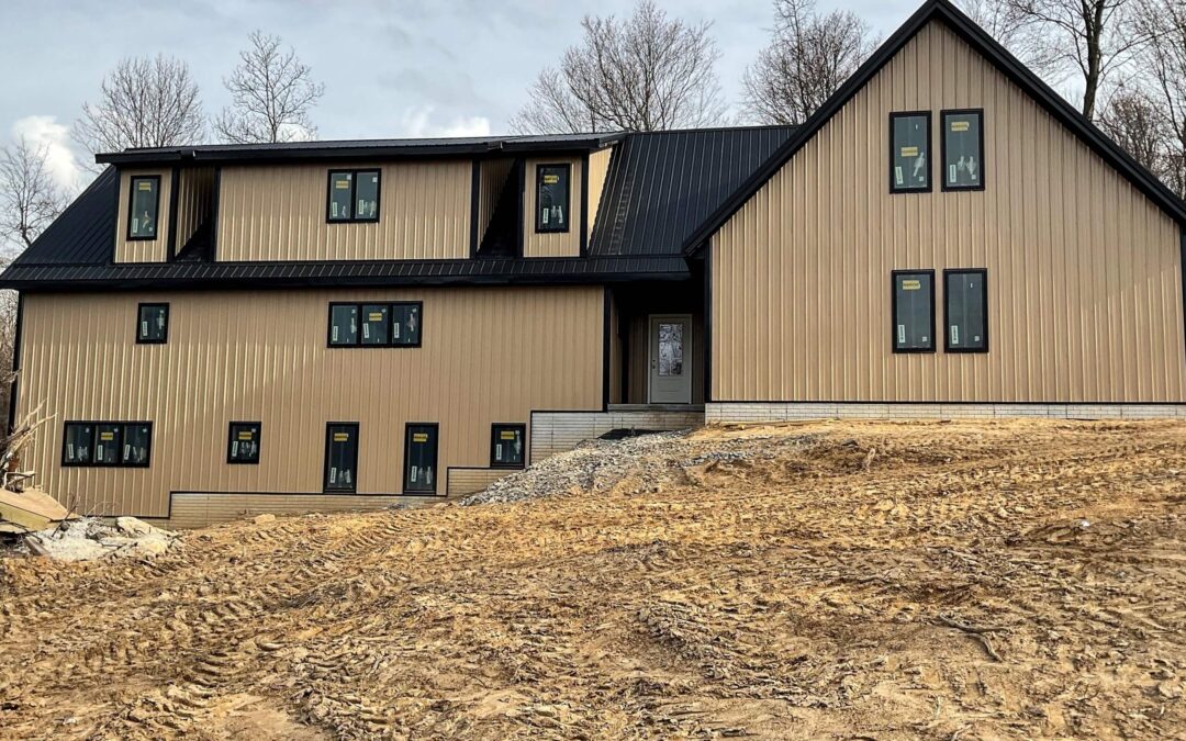 Amish Ridge Metals Mt Perry OH A modern, beige house with black trim and a steep metal roof, set on a bare, dirt-covered lot with trees in the background.